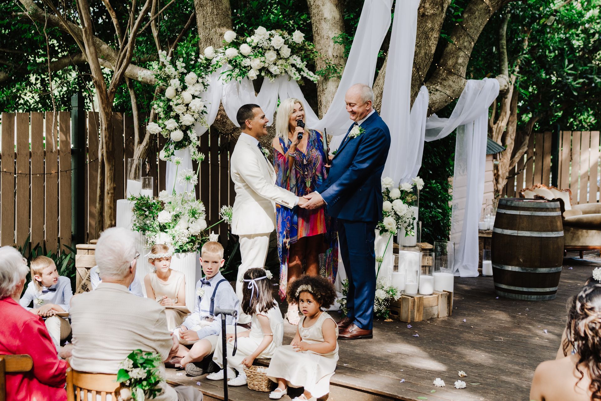 Wedding ceremony: Two grooms holding hands under floral arch, guests seated, outdoor setting.