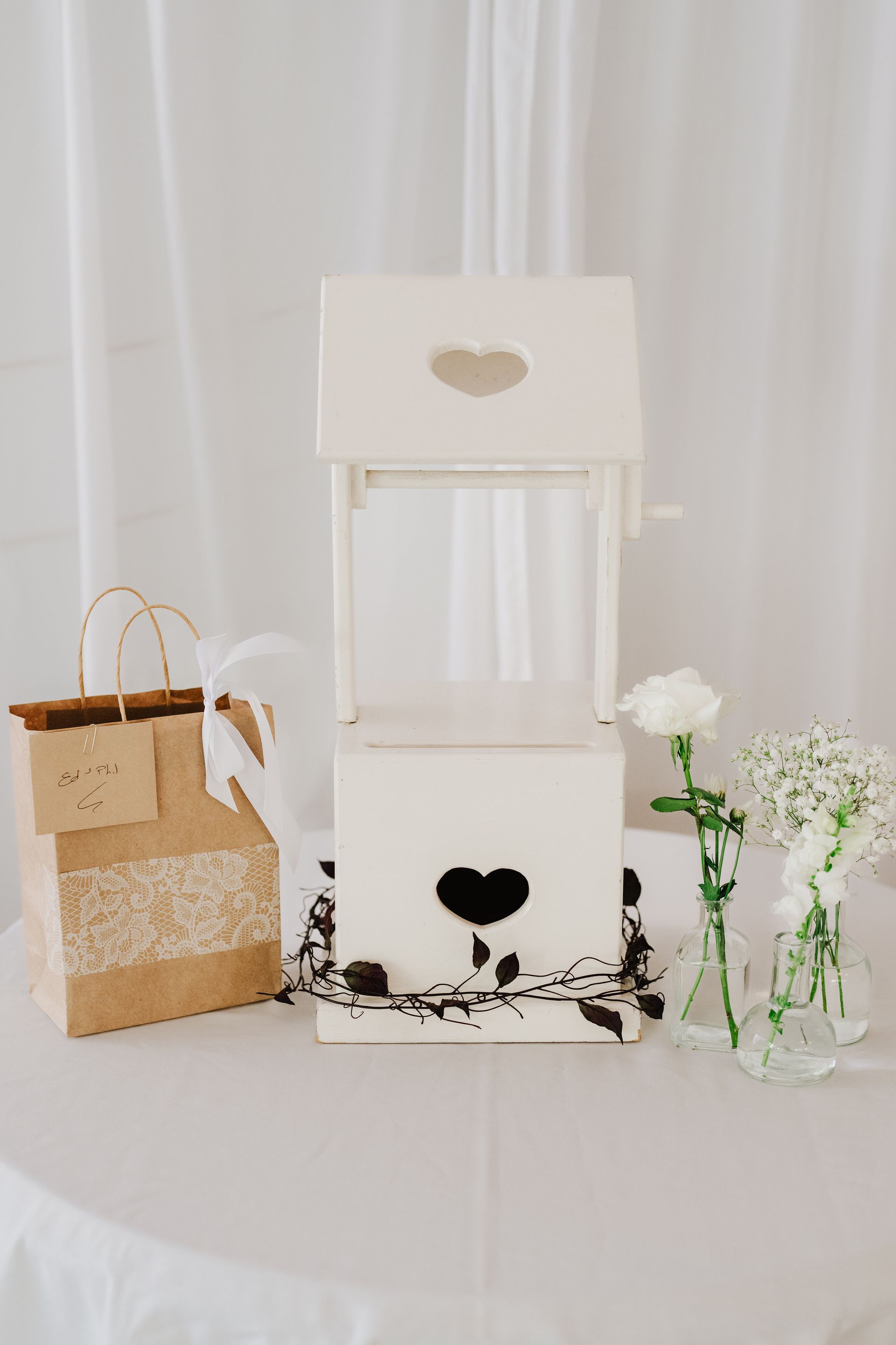 White wishing well with heart cutouts, gift bag, and floral decorations on a white tablecloth.