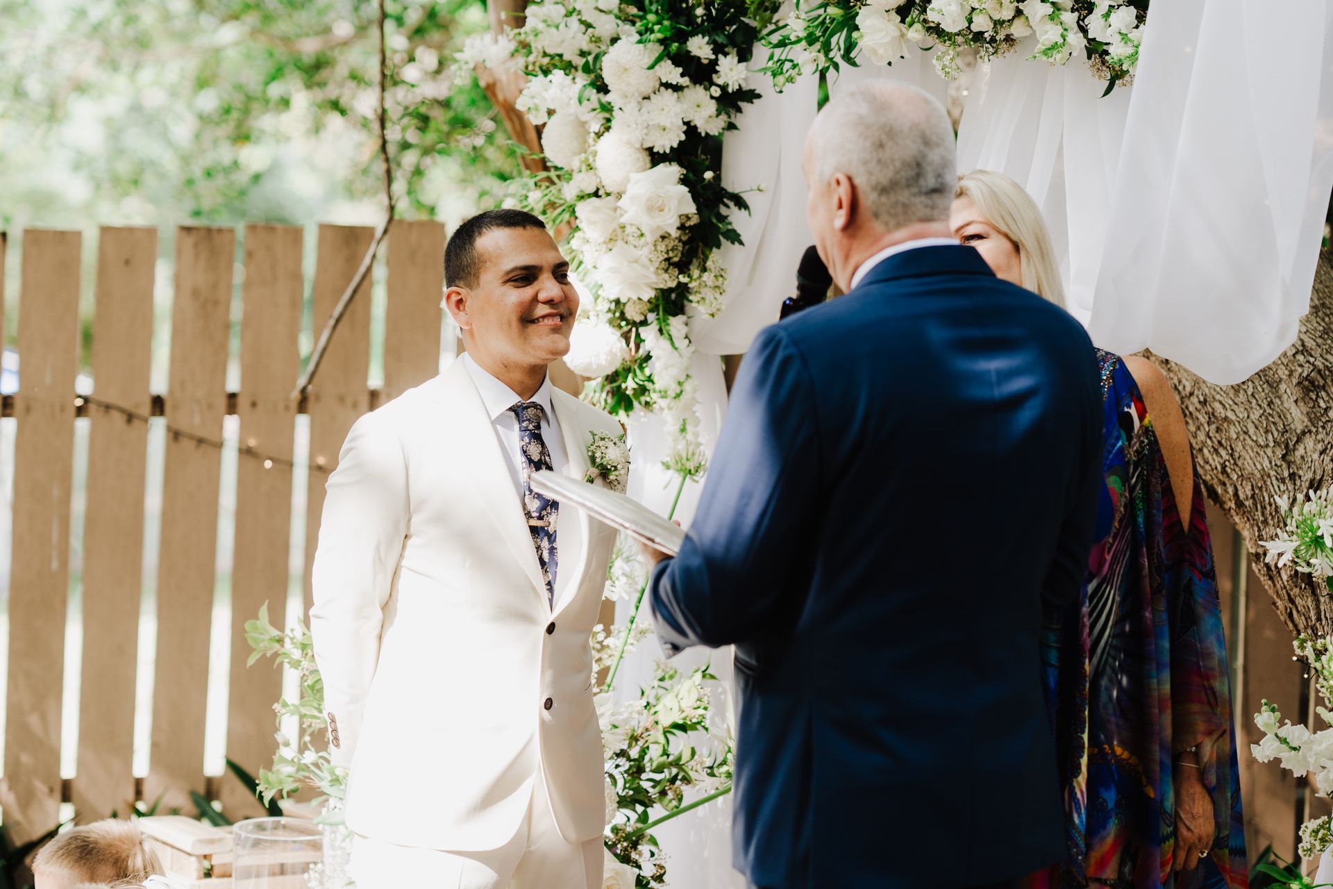 Groom smiles at officiant at outdoor wedding. White flowers and wooden fence in the background.