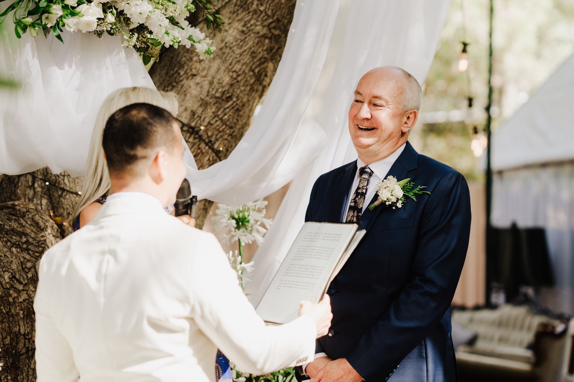 A man in a blue suit smiles at a person holding a document during an outdoor wedding.