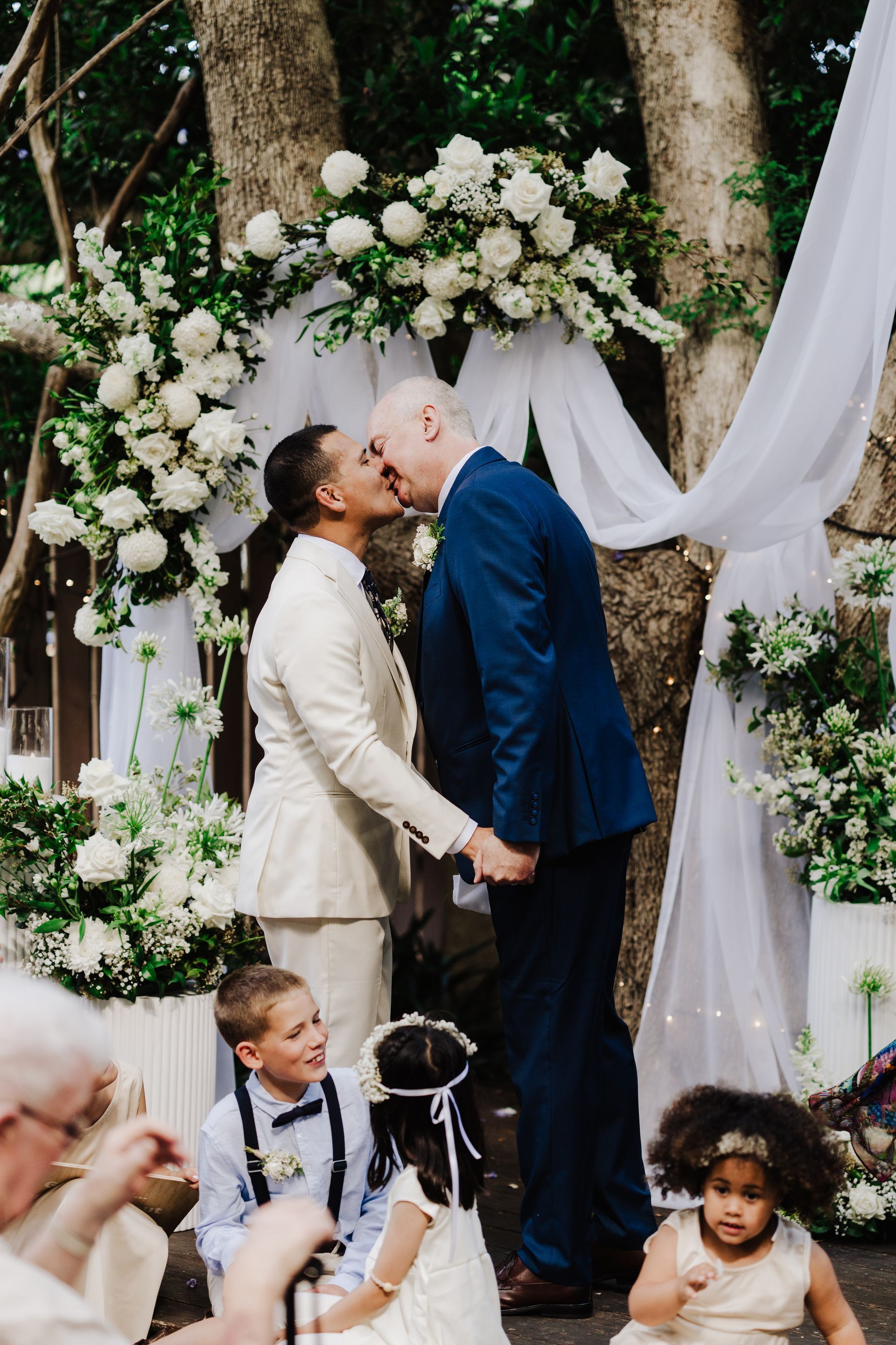 Two men kissing at an outdoor wedding. White floral arch, holding hands, surrounded by guests, and children.
