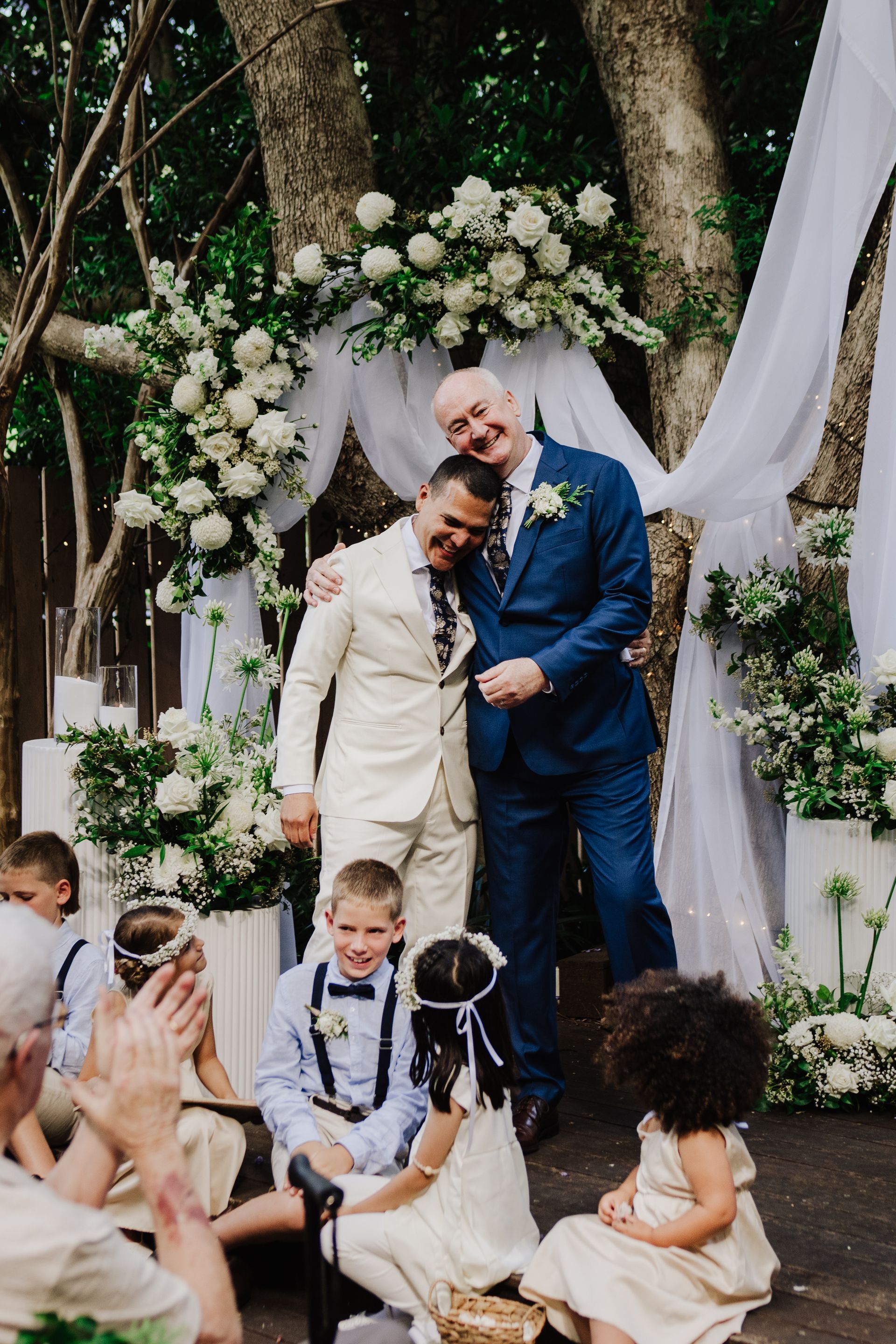 Two men embrace at a wedding altar decorated with white flowers. Children sit in front, smiling.