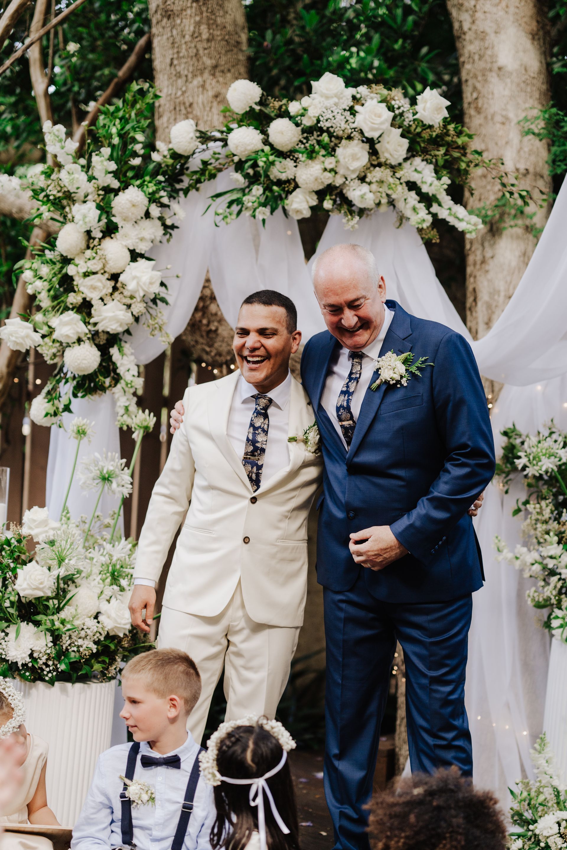 Two men smiling, in suits, at an outdoor wedding ceremony with floral arch, child in foreground.