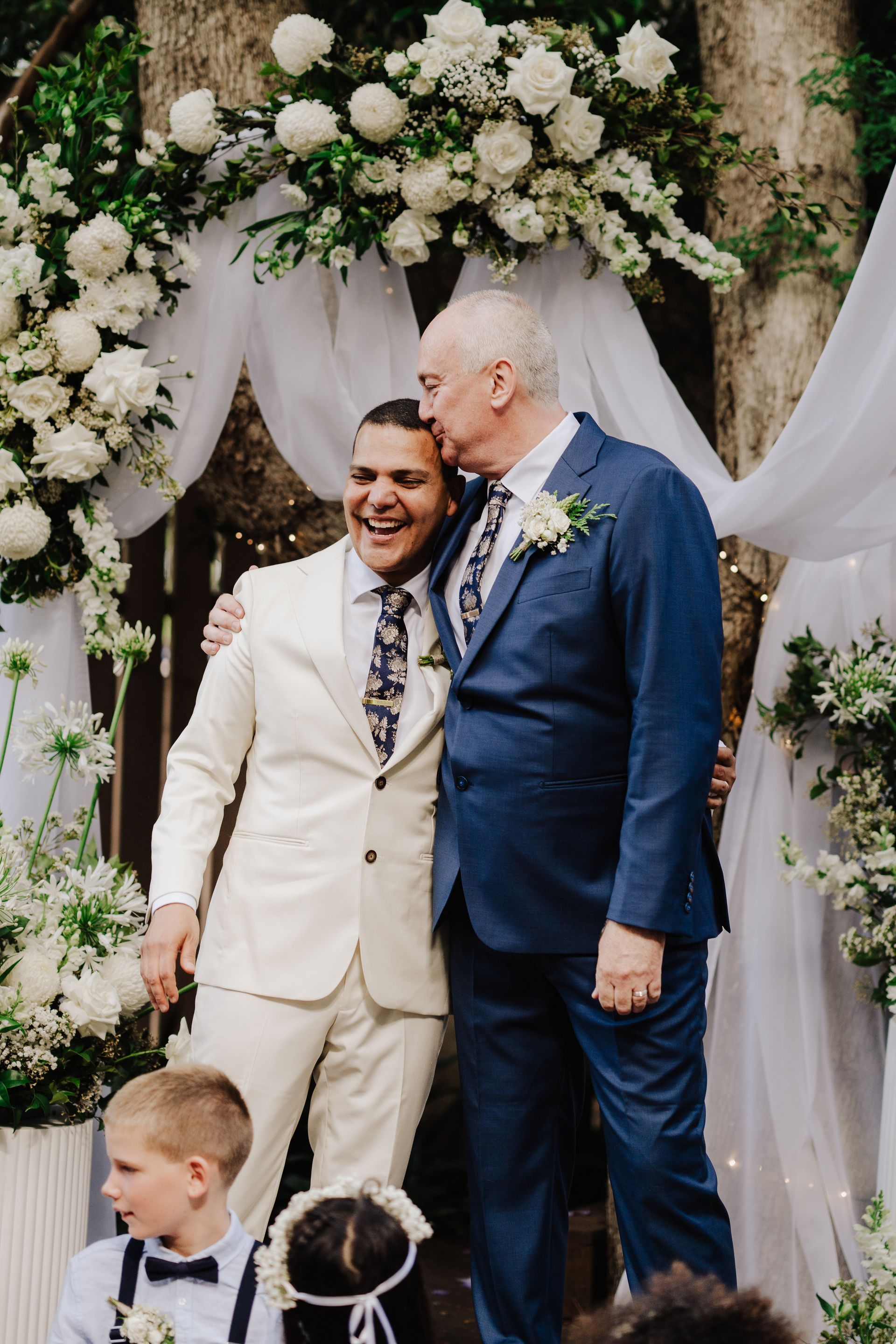 Man in cream suit smiles, embraced by man in blue suit kissing his head, at outdoor wedding with floral arch.