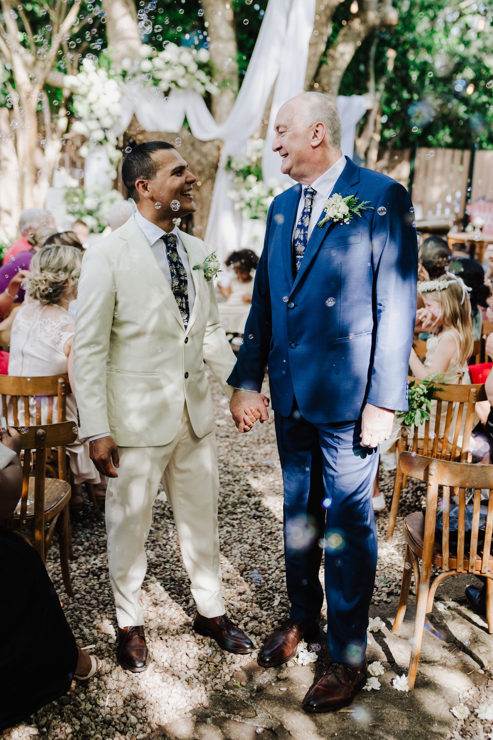 Two grooms holding hands, smiling at each other during an outdoor wedding ceremony. One wears cream suit, the other blue.