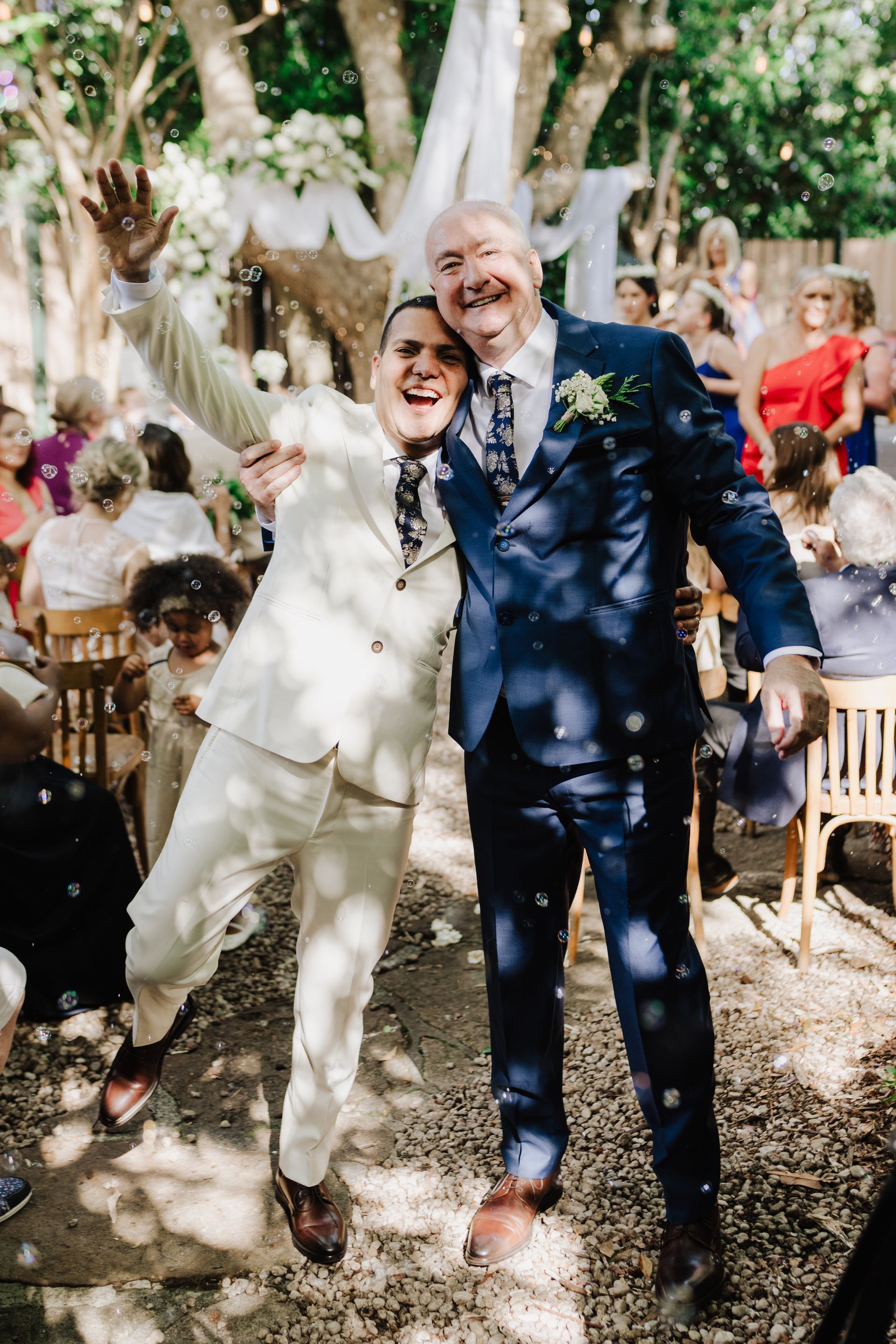 Two men in suits celebrate at an outdoor wedding, smiling, bubbles floating.