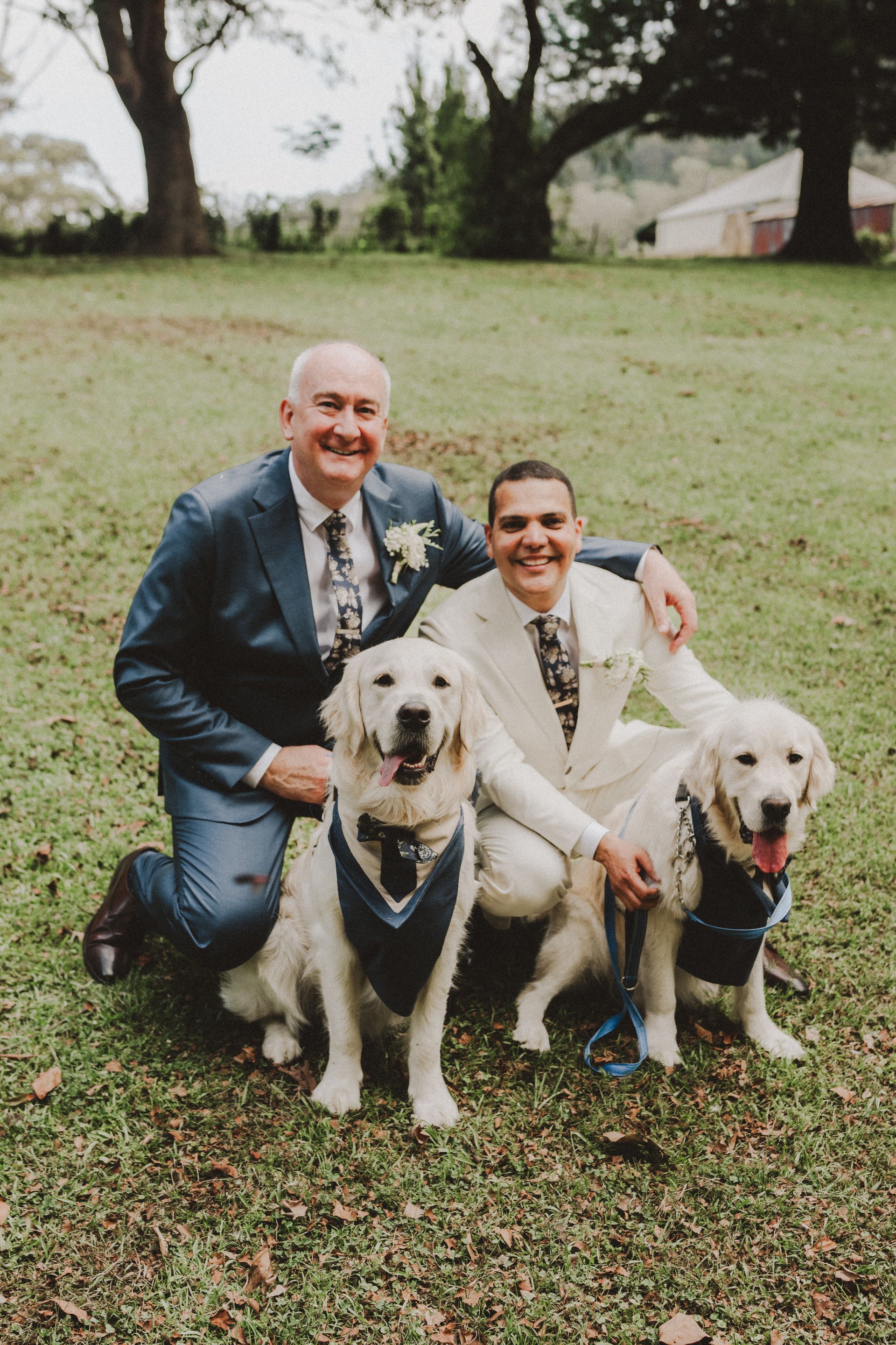 Two men, each with a Golden Retriever, pose outdoors. Men wear suits, dogs sport navy bandanas.