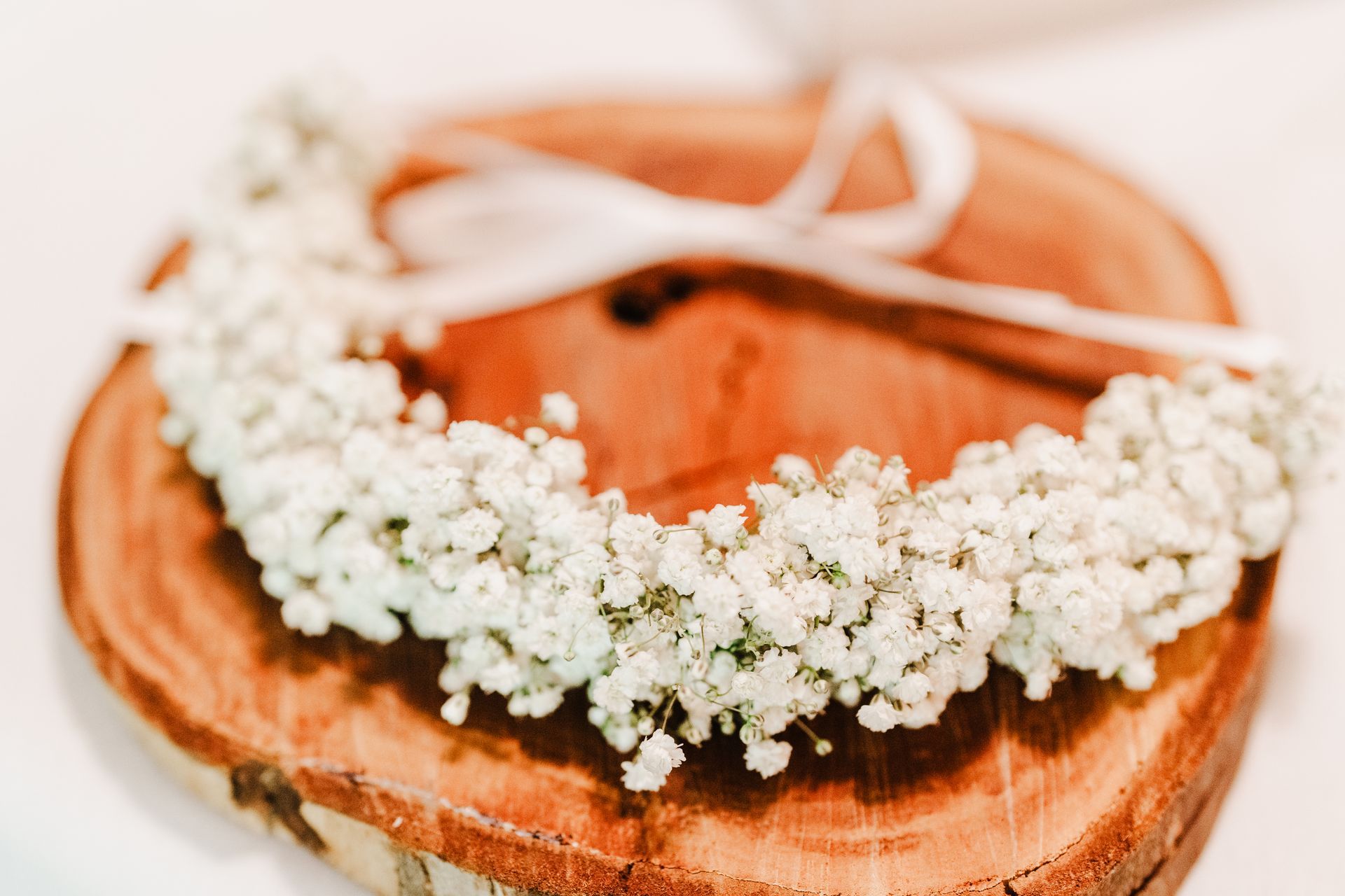 A flower crown made of tiny white flowers rests on a wooden slice, with a white ribbon.