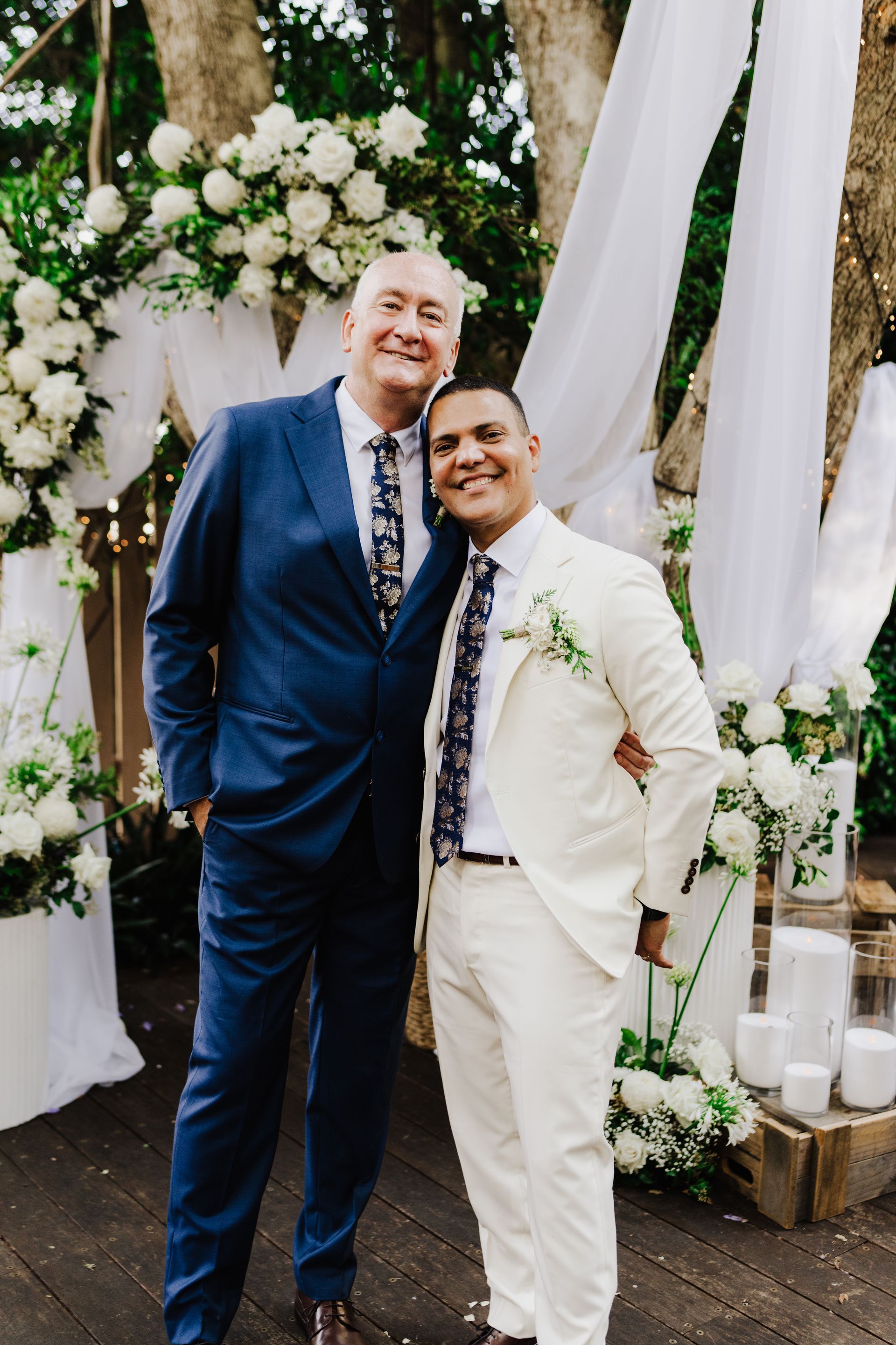 Two men in suits smile, posed near a flower-covered archway during an outdoor wedding.