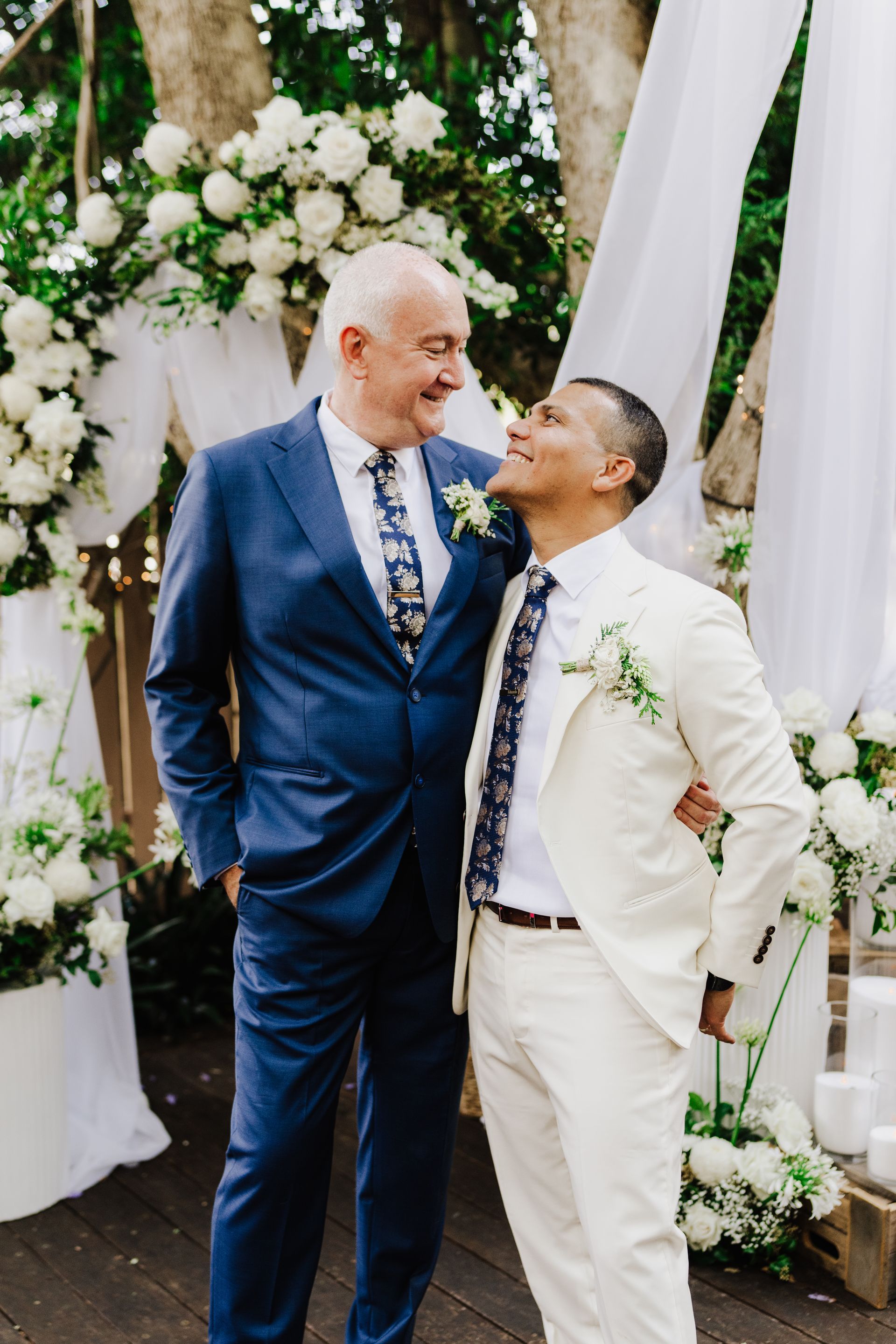 Two men in suits, smiling and embracing at an outdoor wedding.