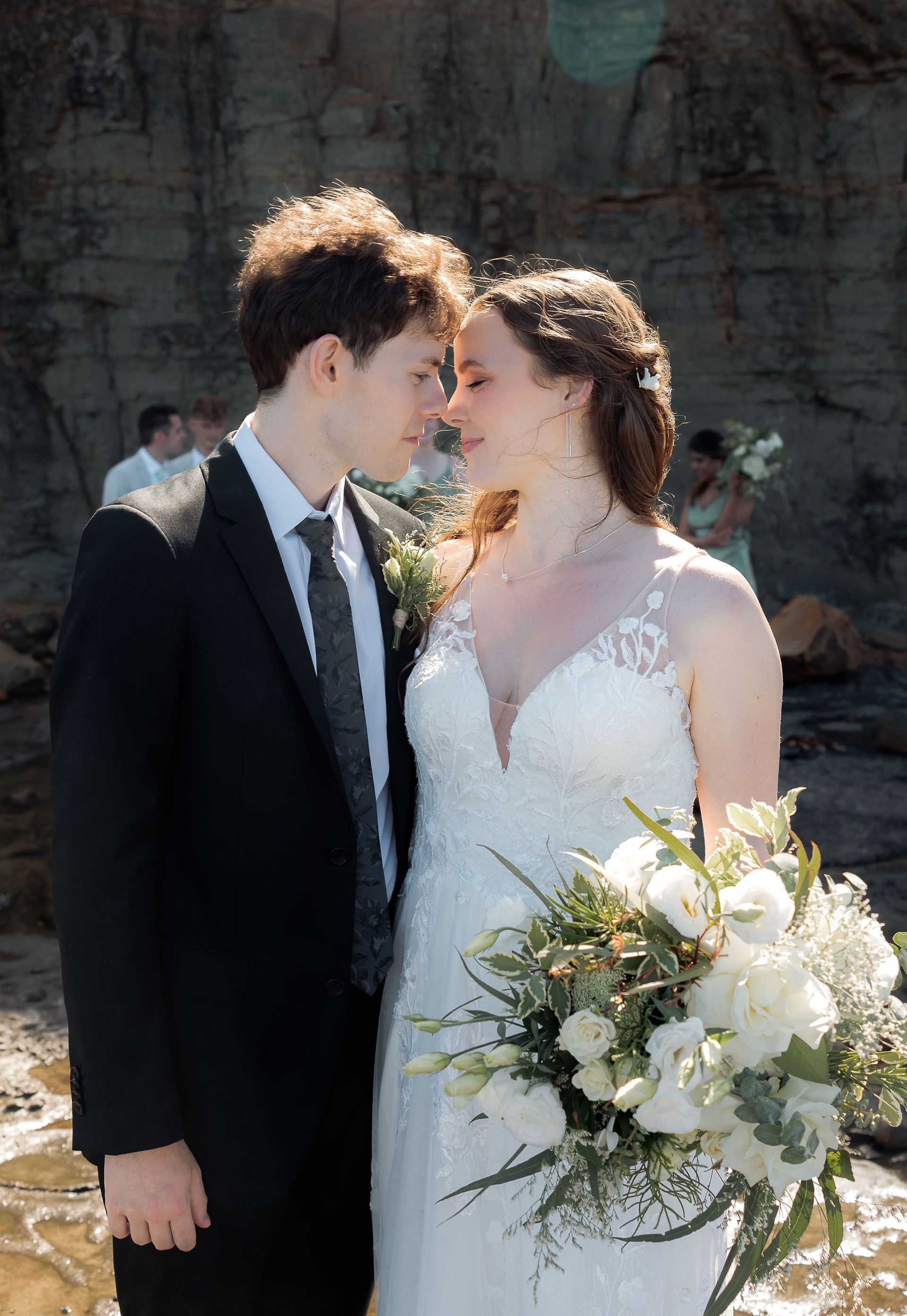 Bride wearing white dress and groom in black suit embrace on a rocky shore. 