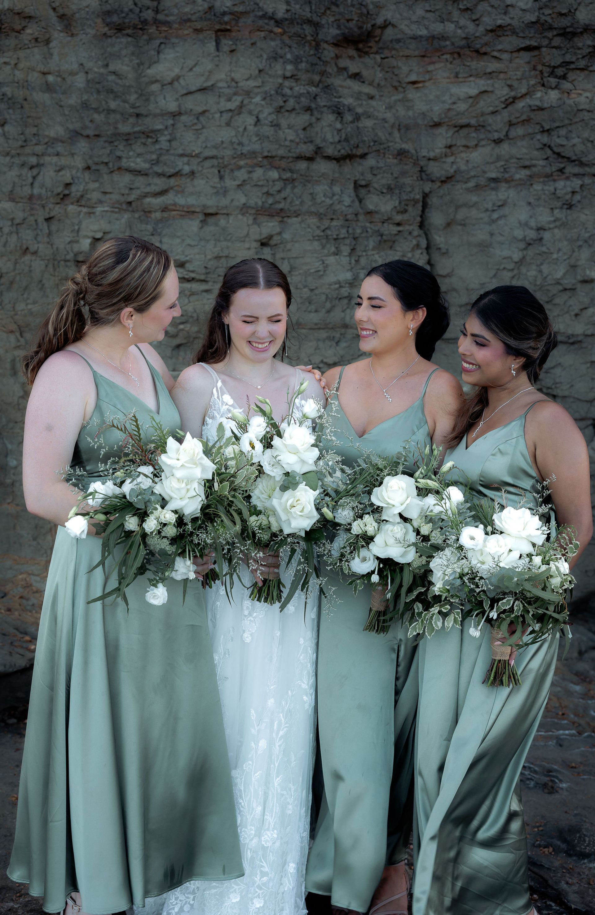 Bride with bridesmaids in sage green dresses, holding bouquets, laughing against a rock wall.