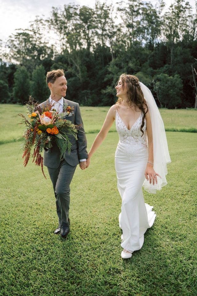 Newlyweds walking, holding hands in a field, the bride in a white dress, groom in a suit, smiling.