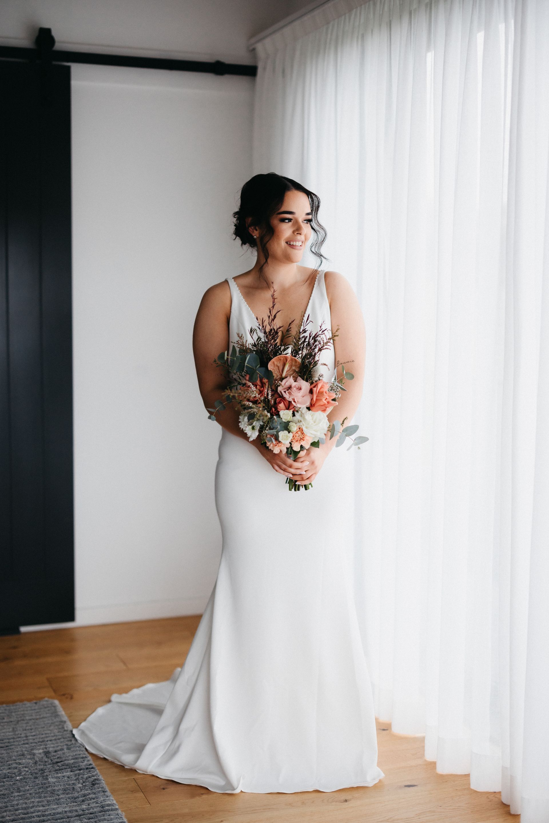 Bride in white gown holds bouquet, smiles near a sheer curtain.