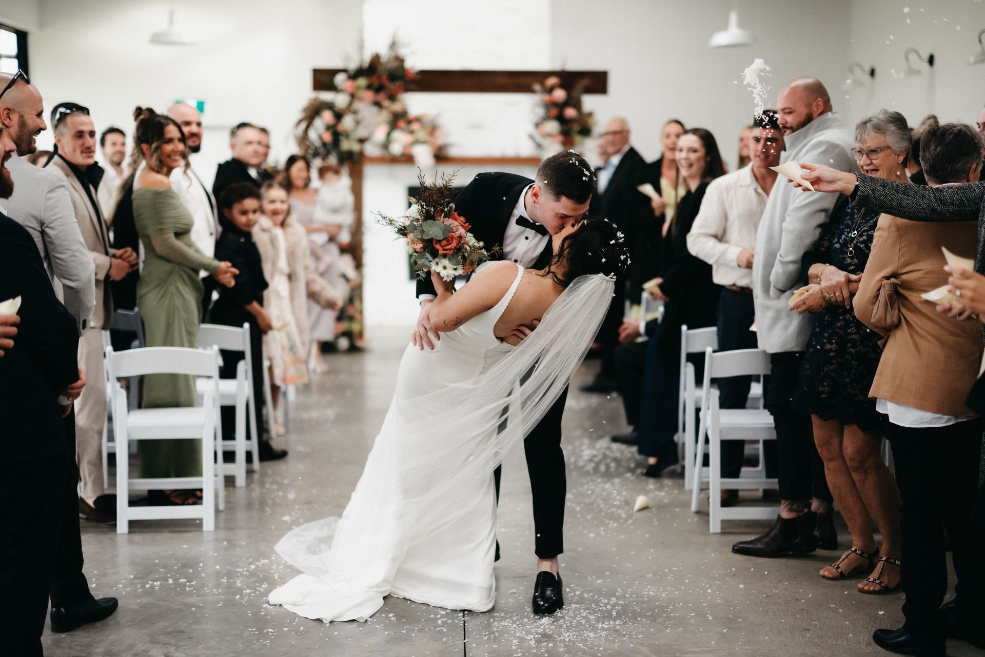 Newlyweds kiss, dipping the bride, while guests cheer and confetti falls at a wedding ceremony.