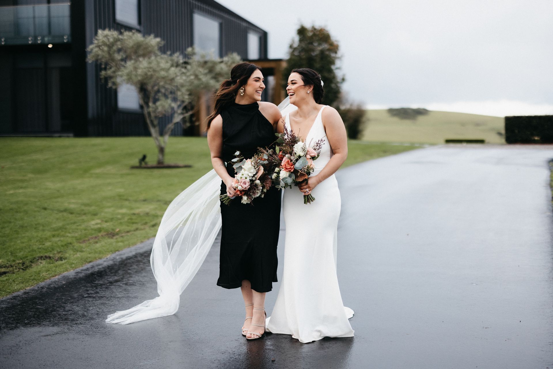 Two women, one in a black dress with a veil, the other in a white dress, walk arm in arm on a wet path, smiling.