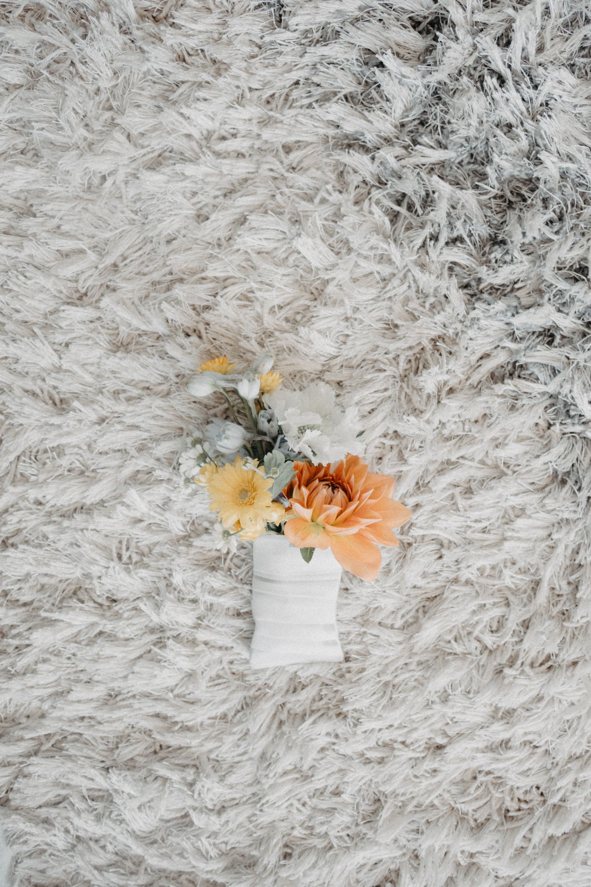 Bouquet of yellow and orange flowers wrapped in white ribbon, resting on a plush, white rug.