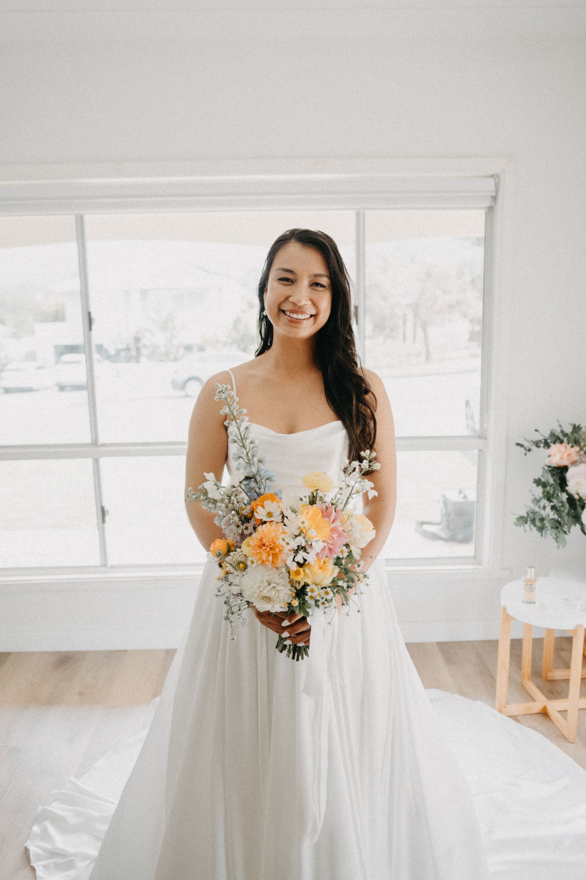 Woman in a white wedding dress smiling, holding a floral bouquet, standing by a window.