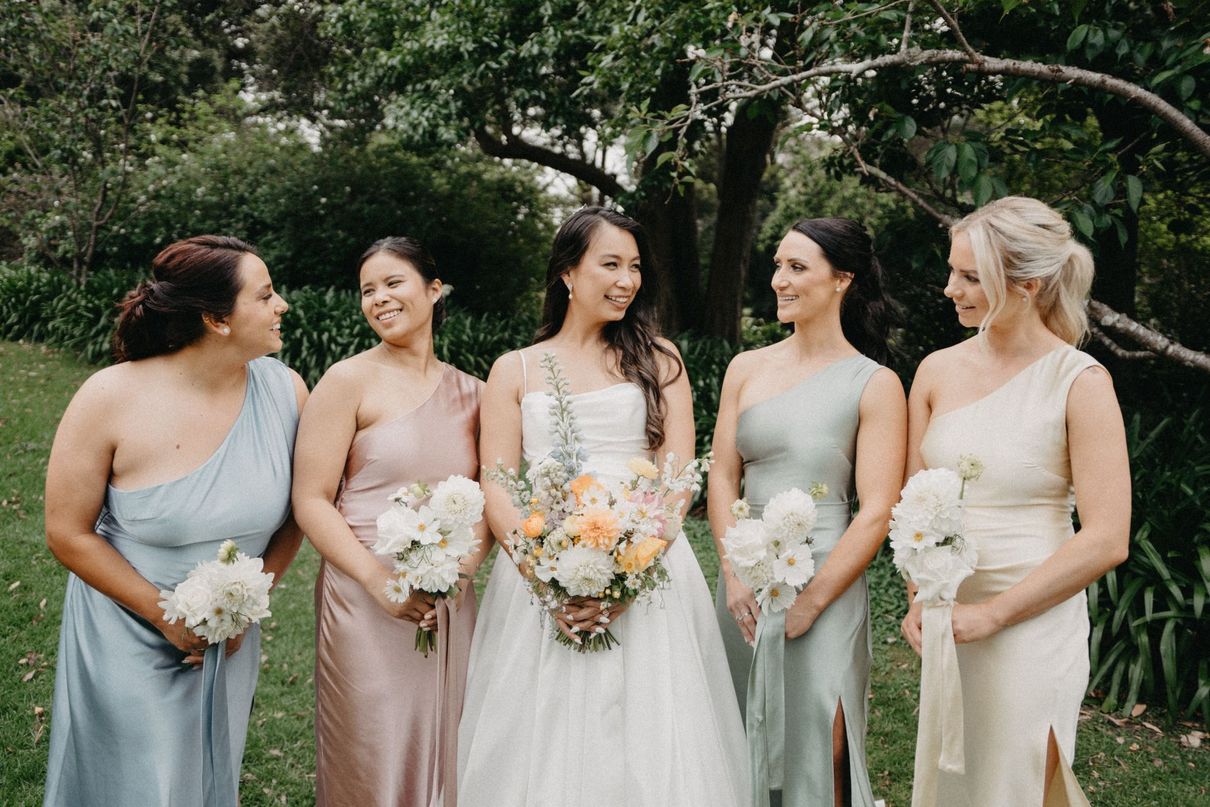 Bride and bridesmaids pose outdoors. Bride in white, bridesmaids in gray dresses, holding flowers.