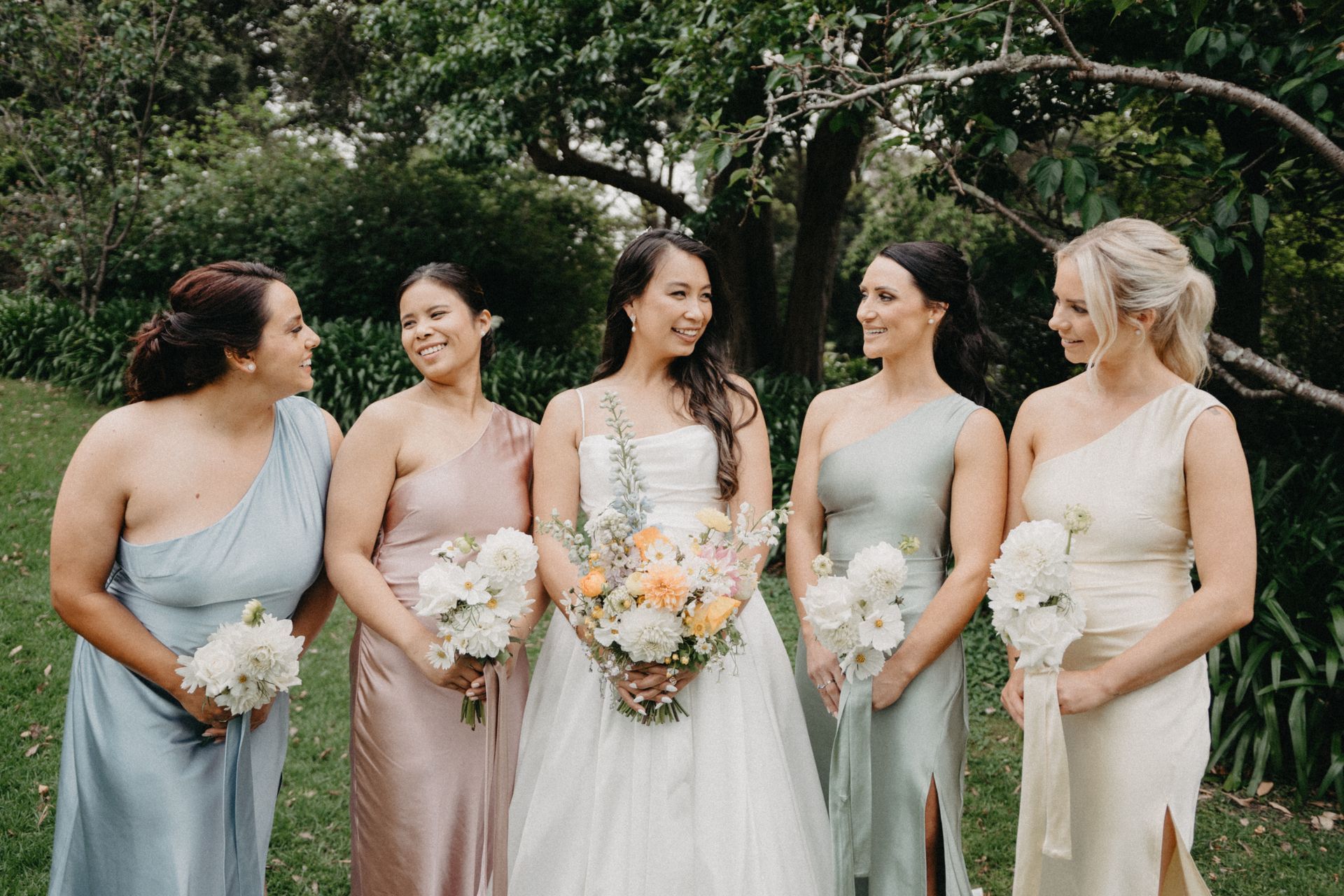 Bride and bridesmaids in colorful gowns with bouquets, outdoors.