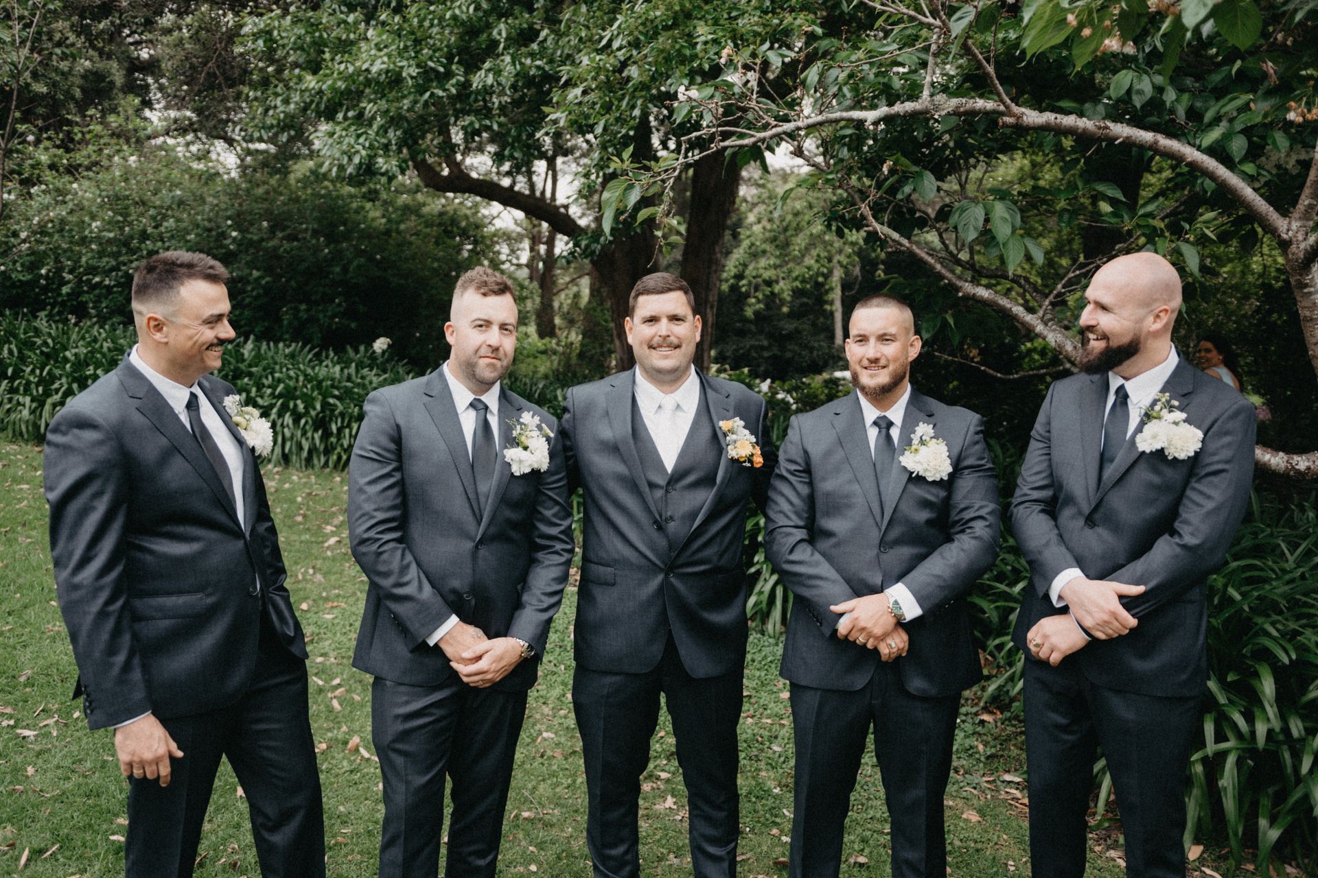 Five men in dark gray suits pose outdoors in front of greenery.