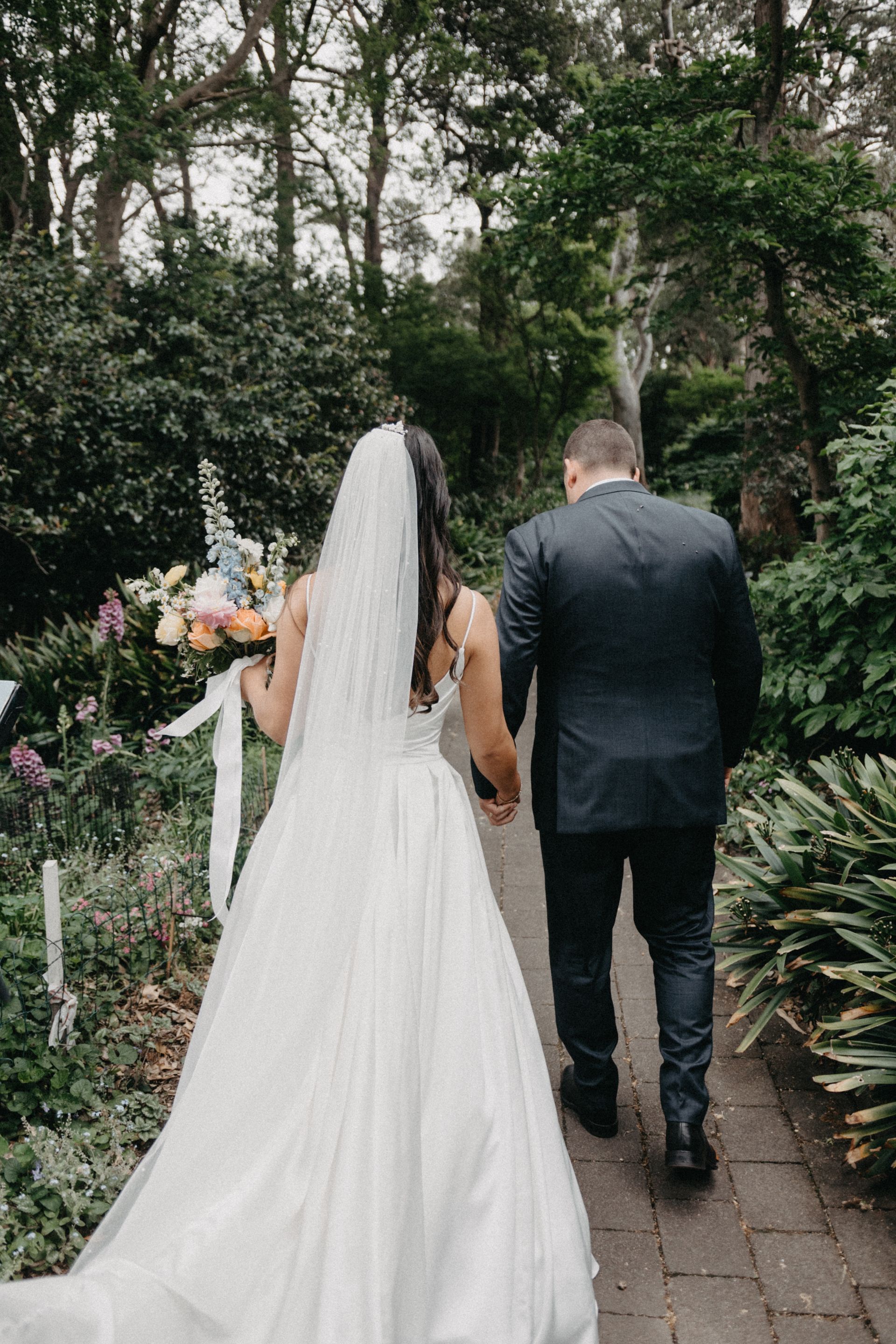 Bride and groom walking away, holding hands, in a garden. She wears a white dress with a veil and holds flowers.