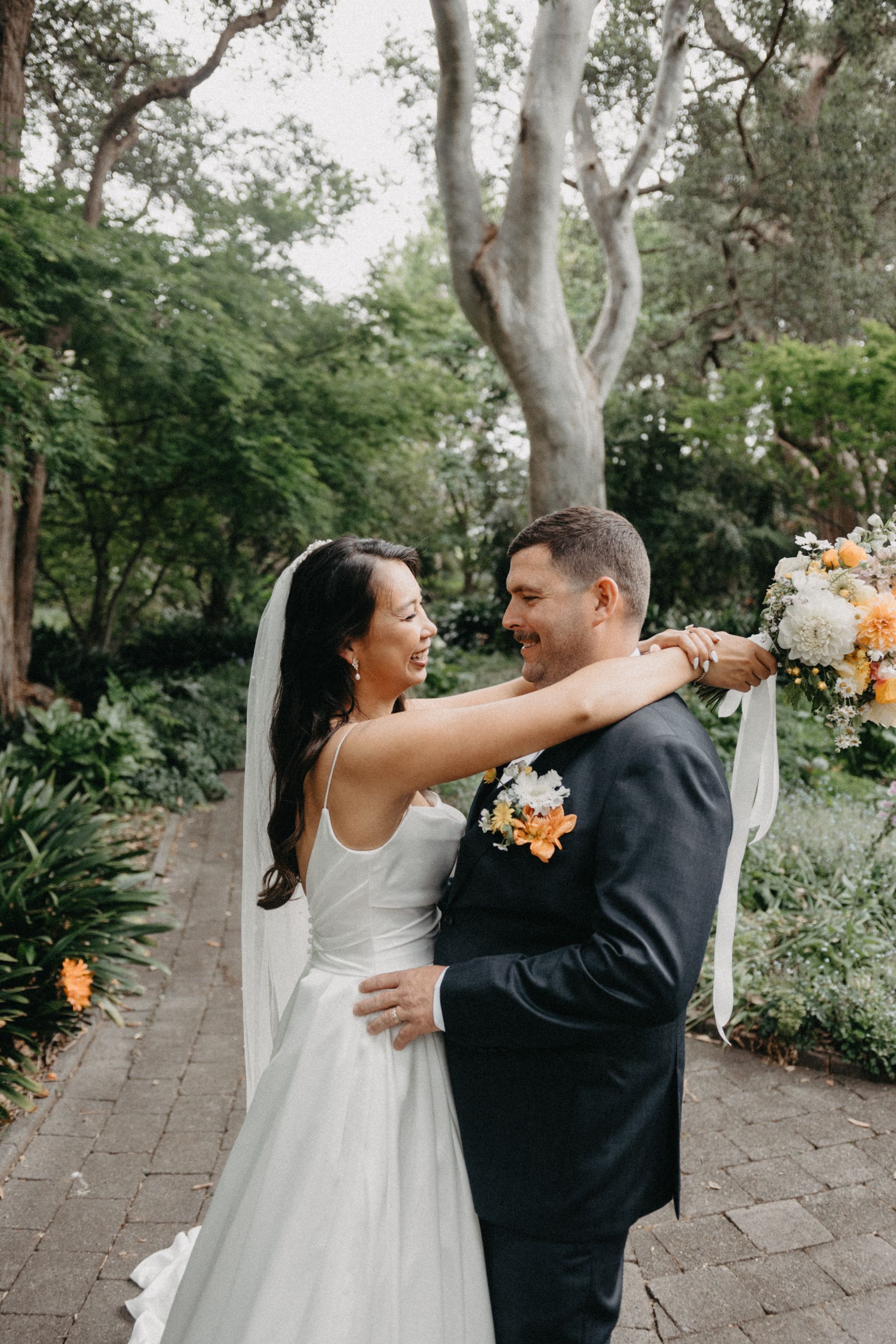 Bride and groom embrace outdoors, smiling at each other. Lush greenery and stone path surround them.