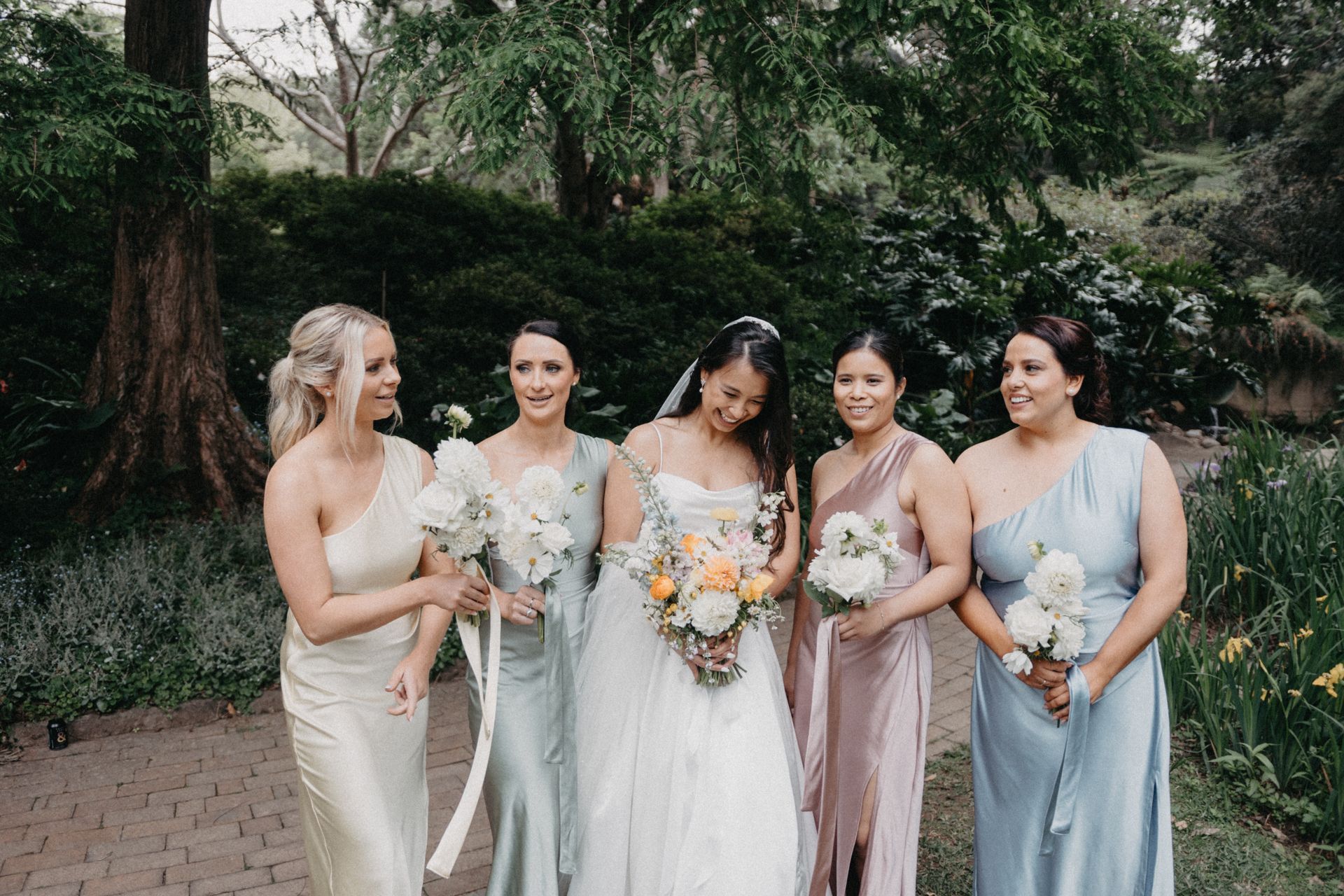 Bride and bridesmaids in a garden, wearing pastel gowns and holding bouquets.