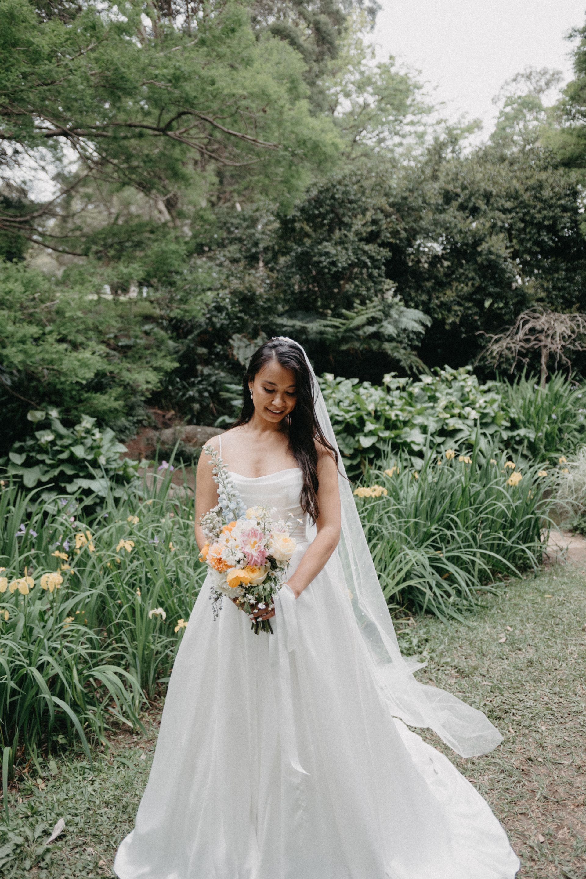 Bride in white dress holding bouquet, outdoors in garden setting.