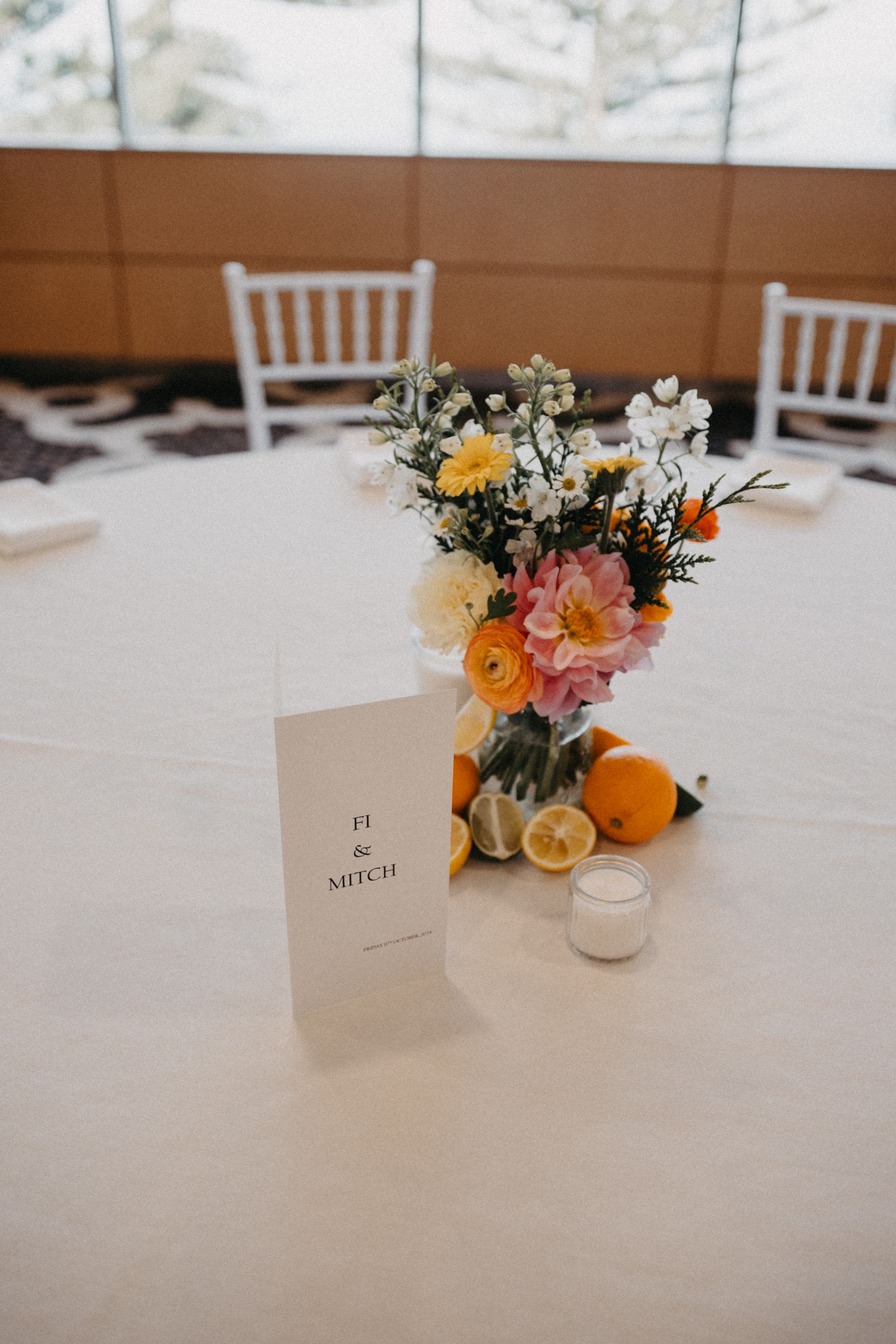 Centerpiece with flowers, fruit, and a sign on a white tablecloth in a bright room.
