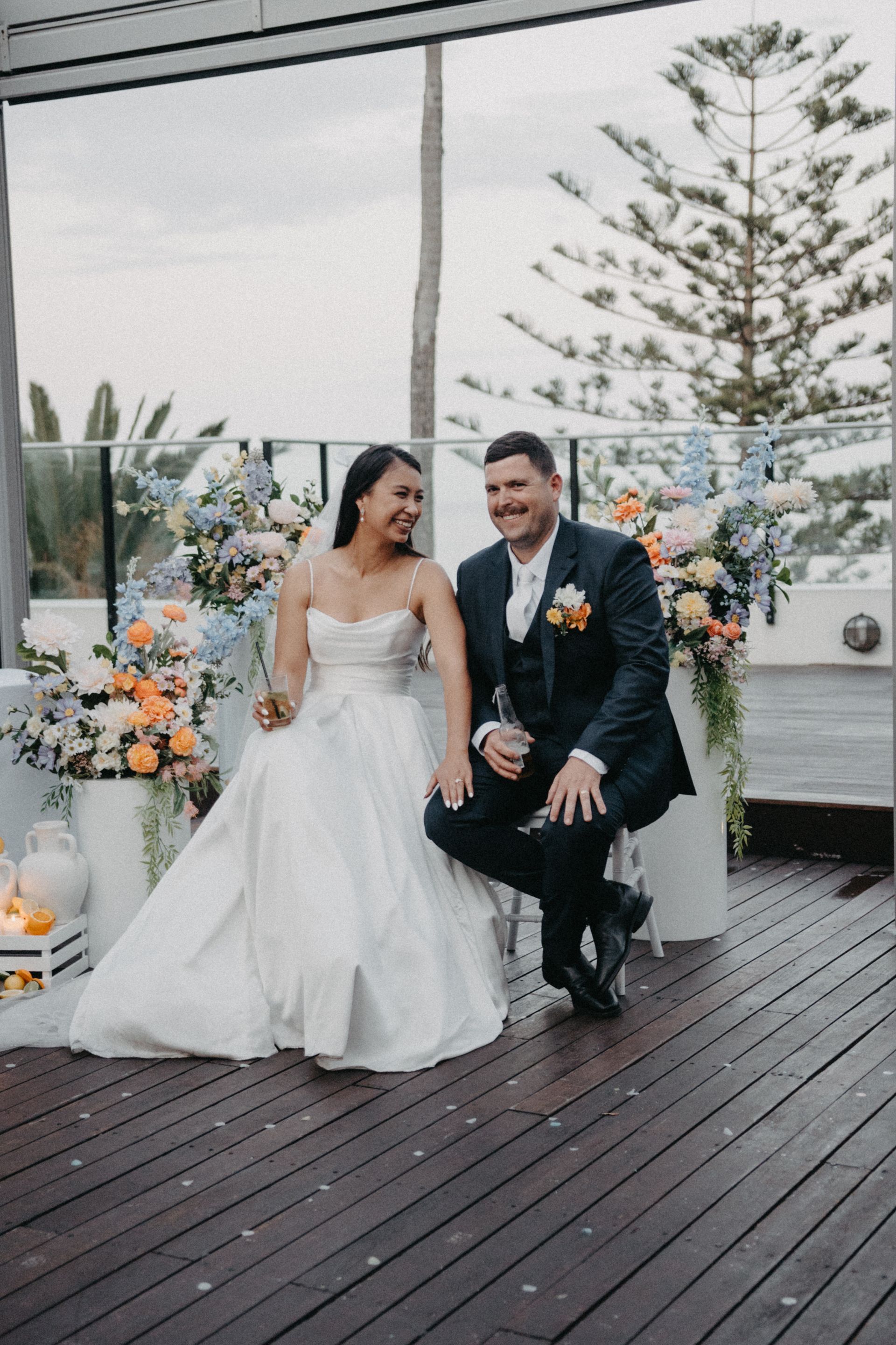 Bride and groom smiling at each other, sitting on a wooden deck, surrounded by floral arrangements.
