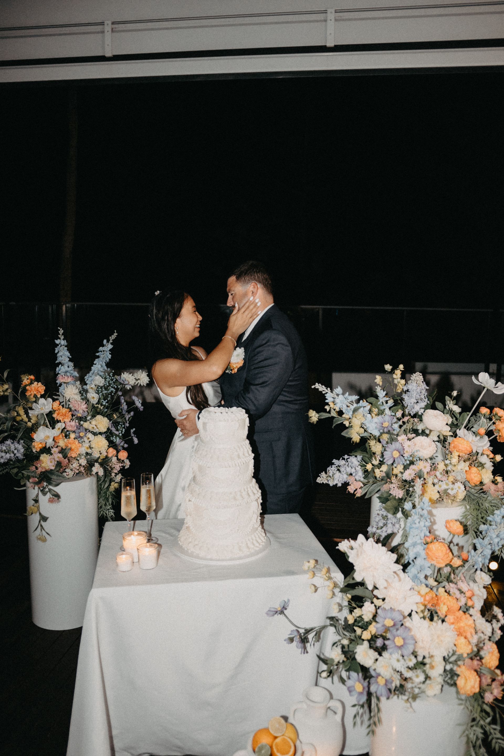 Bride playfully smears cake on groom's face. They stand near a tiered cake.