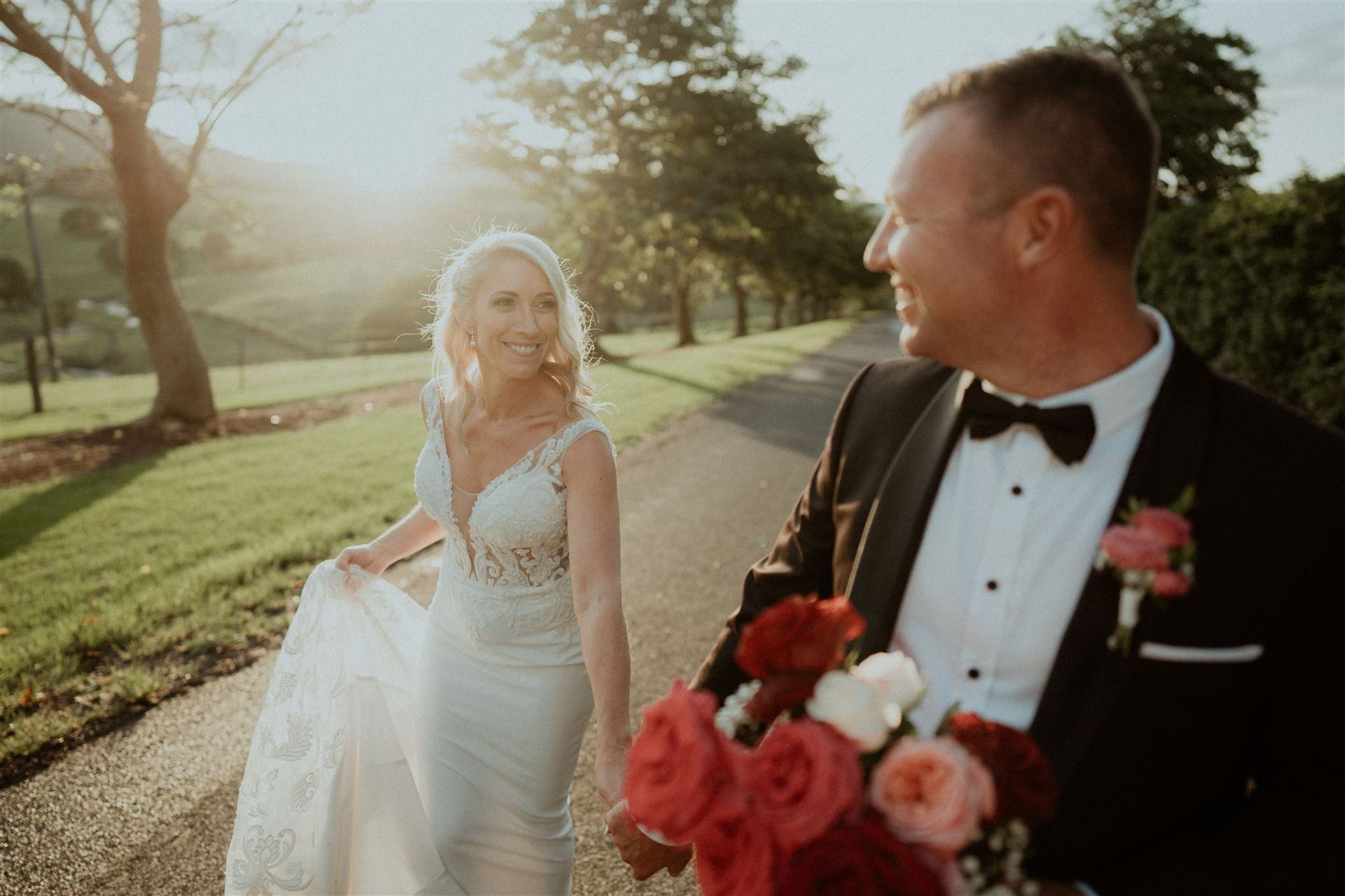 Bride and groom walking on a path, holding hands, smiling. Wedding dress, bouquet, sunny setting.