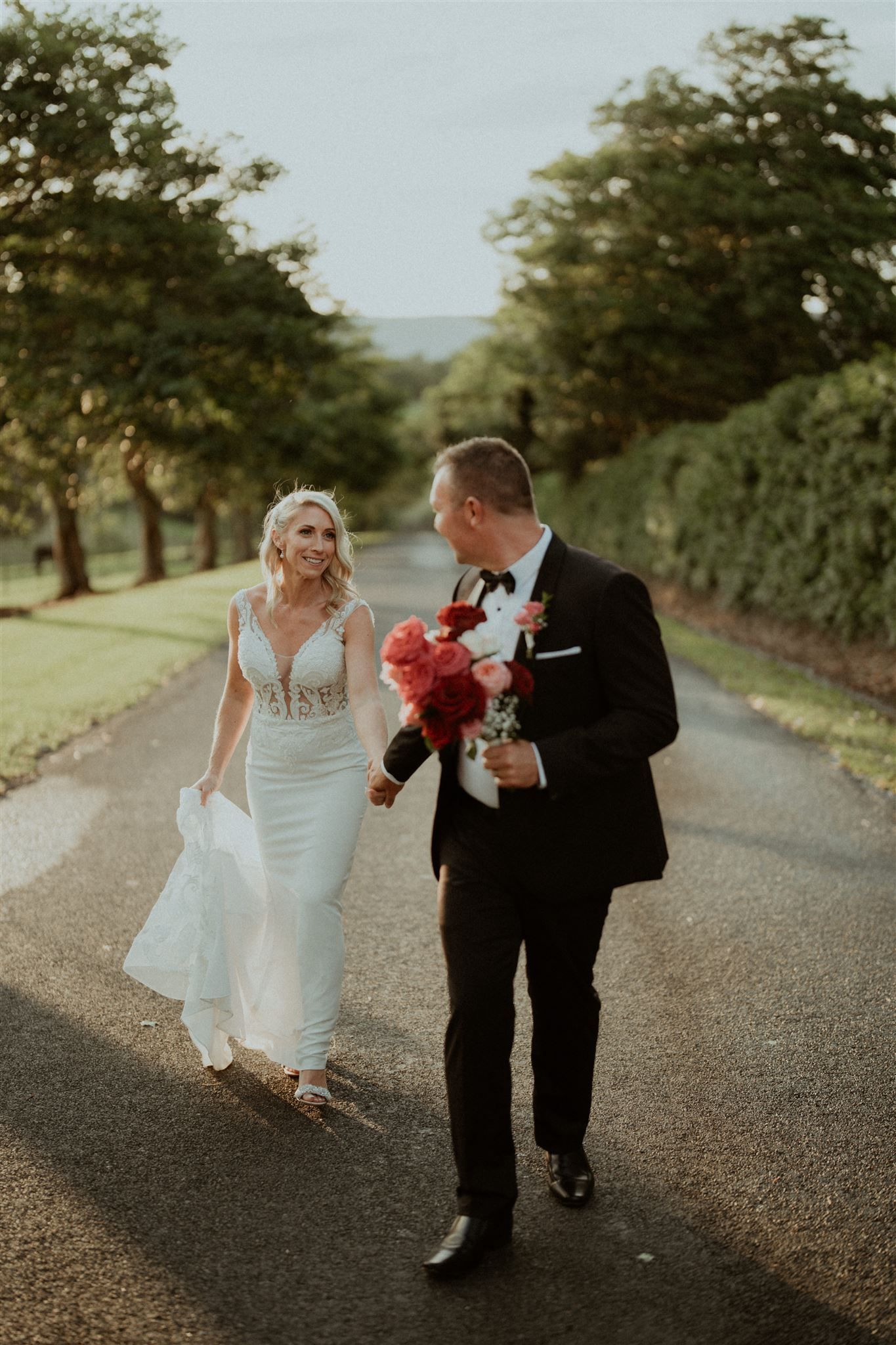 Newlyweds walking on a tree-lined path, bride holding her dress and a bouquet; groom smiles, holding her hand.