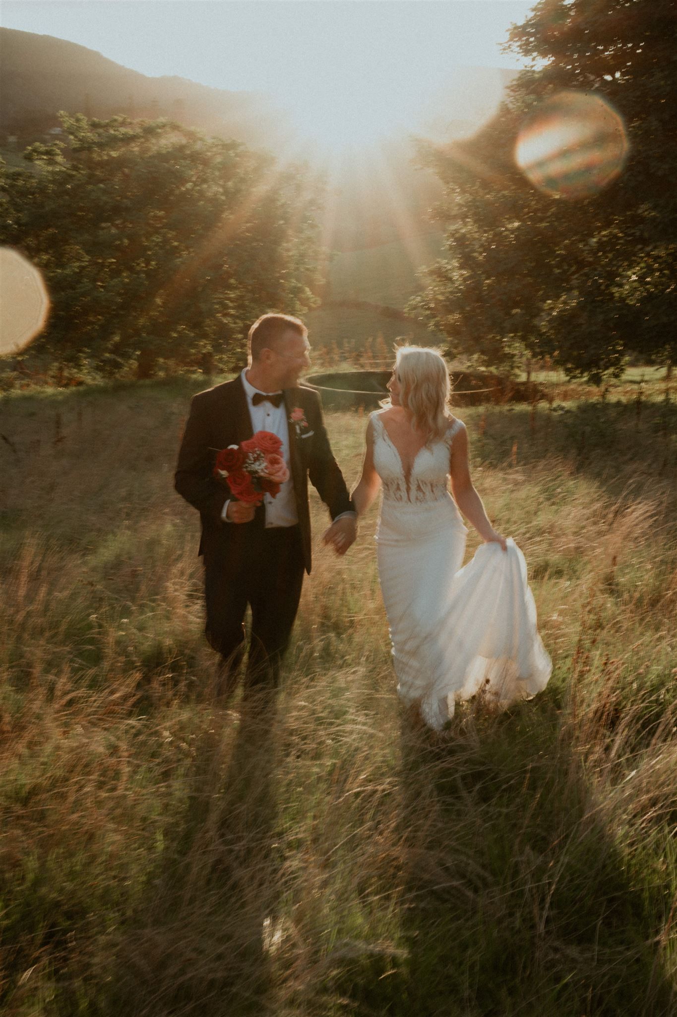 Newlyweds walk hand-in-hand through a sunlit field; bride in white gown, groom in tuxedo, carrying flowers.