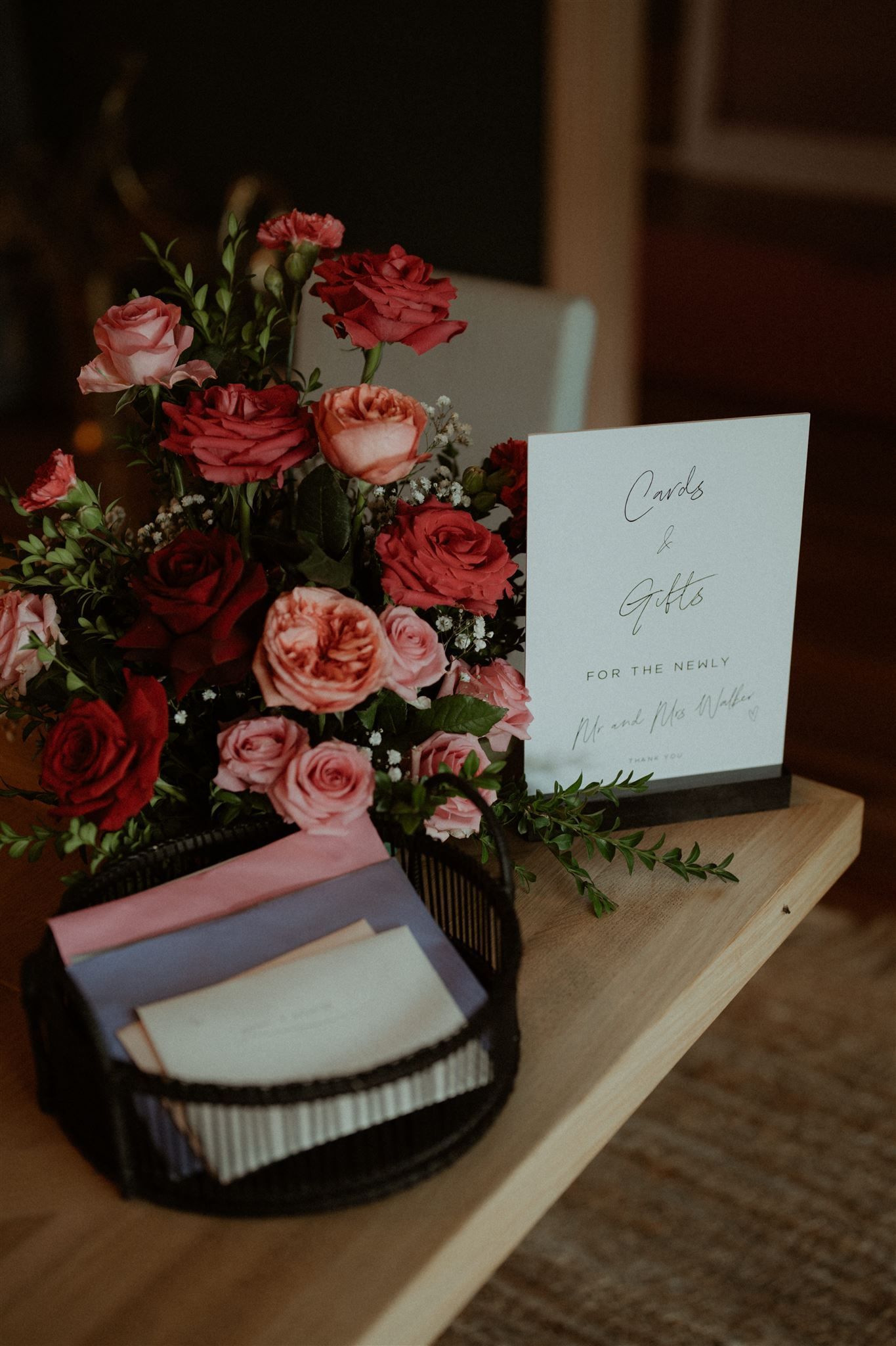 Arrangement of red and pink roses, sign, and basket of cards on a wooden table.