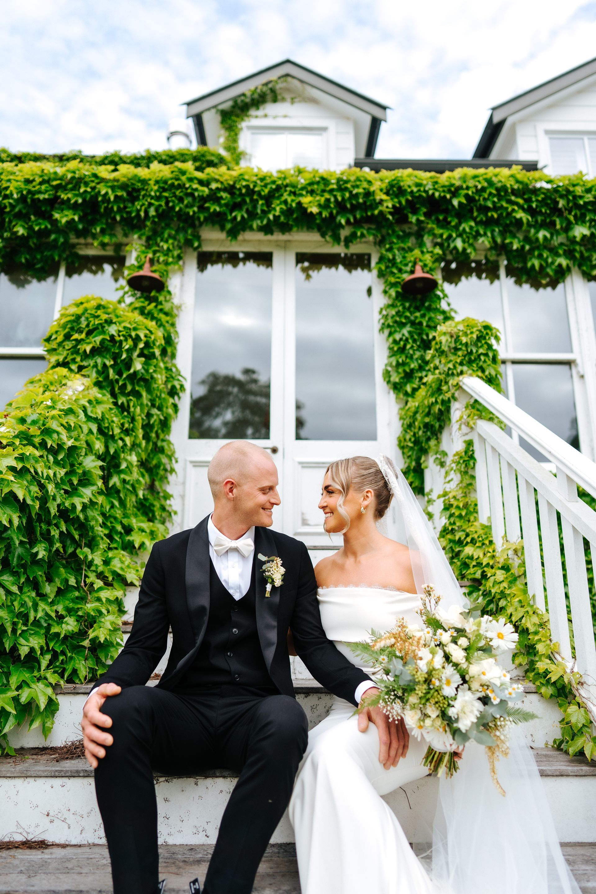 Bride and groom sitting on steps, smiling at each other in front of a white house covered in green vines.