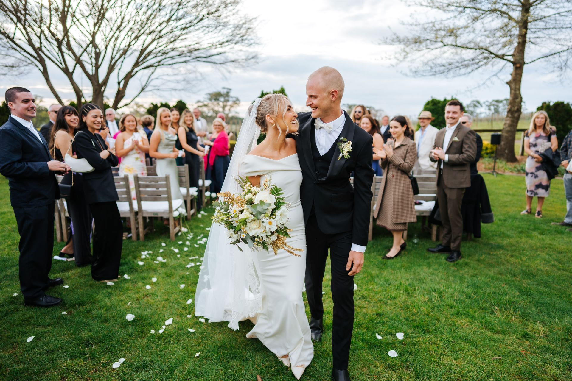 Newlyweds smiling as they walk down aisle lined with cheering guests in outdoor setting.