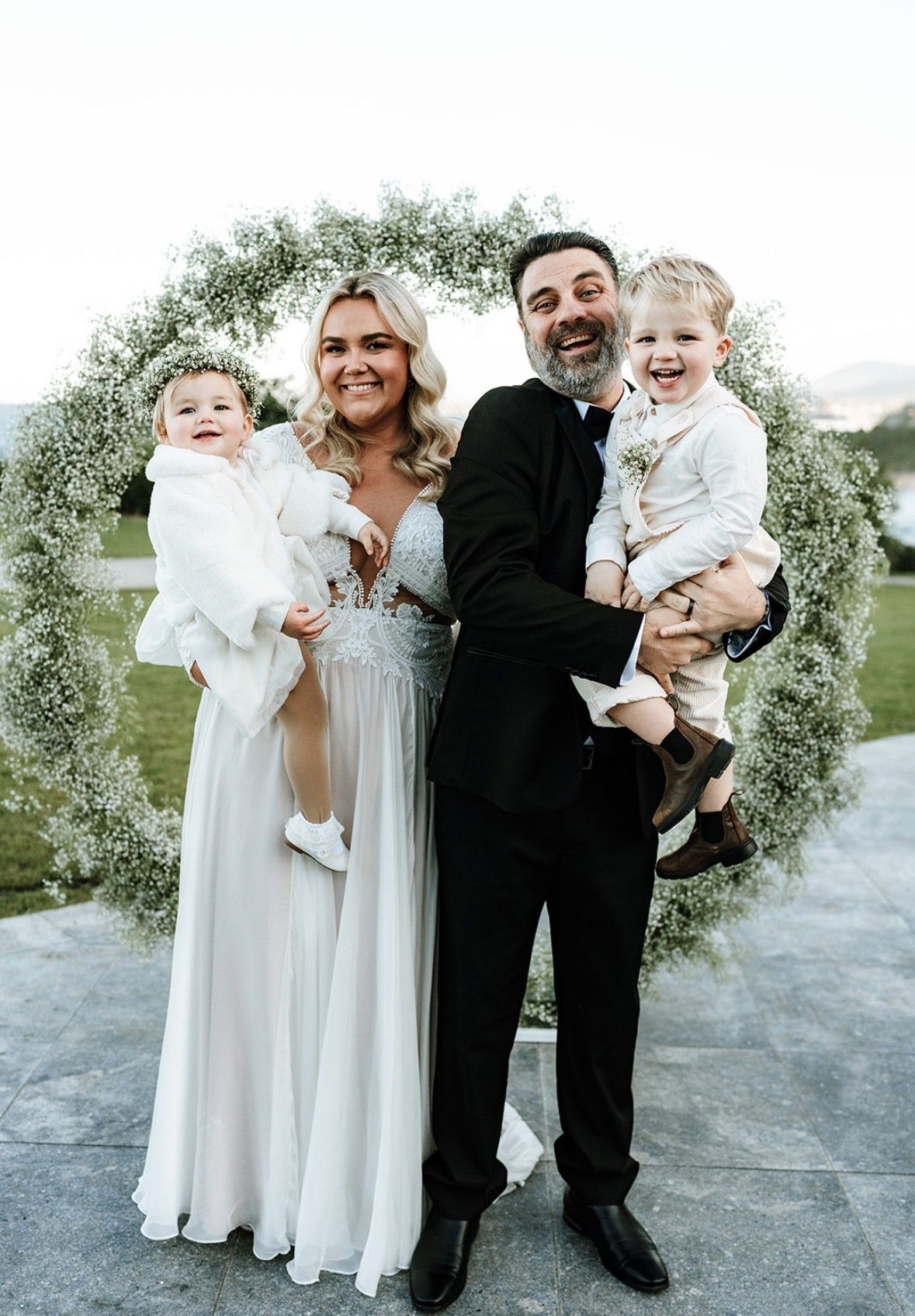 Family poses in front of floral wreath; woman in white gown holds baby, man in suit holds toddler, all smiling.