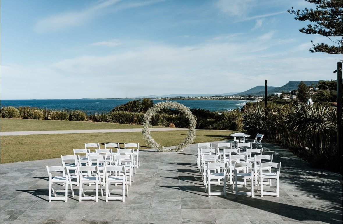 Wedding ceremony setup overlooking the ocean, white chairs, floral arch, blue sky.