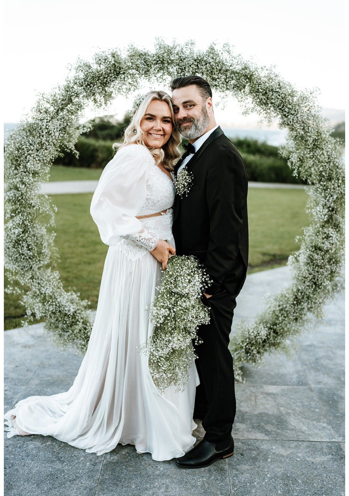 Couple smiling in front of floral archway. Bride in white dress holds bouquet. Groom in black suit.