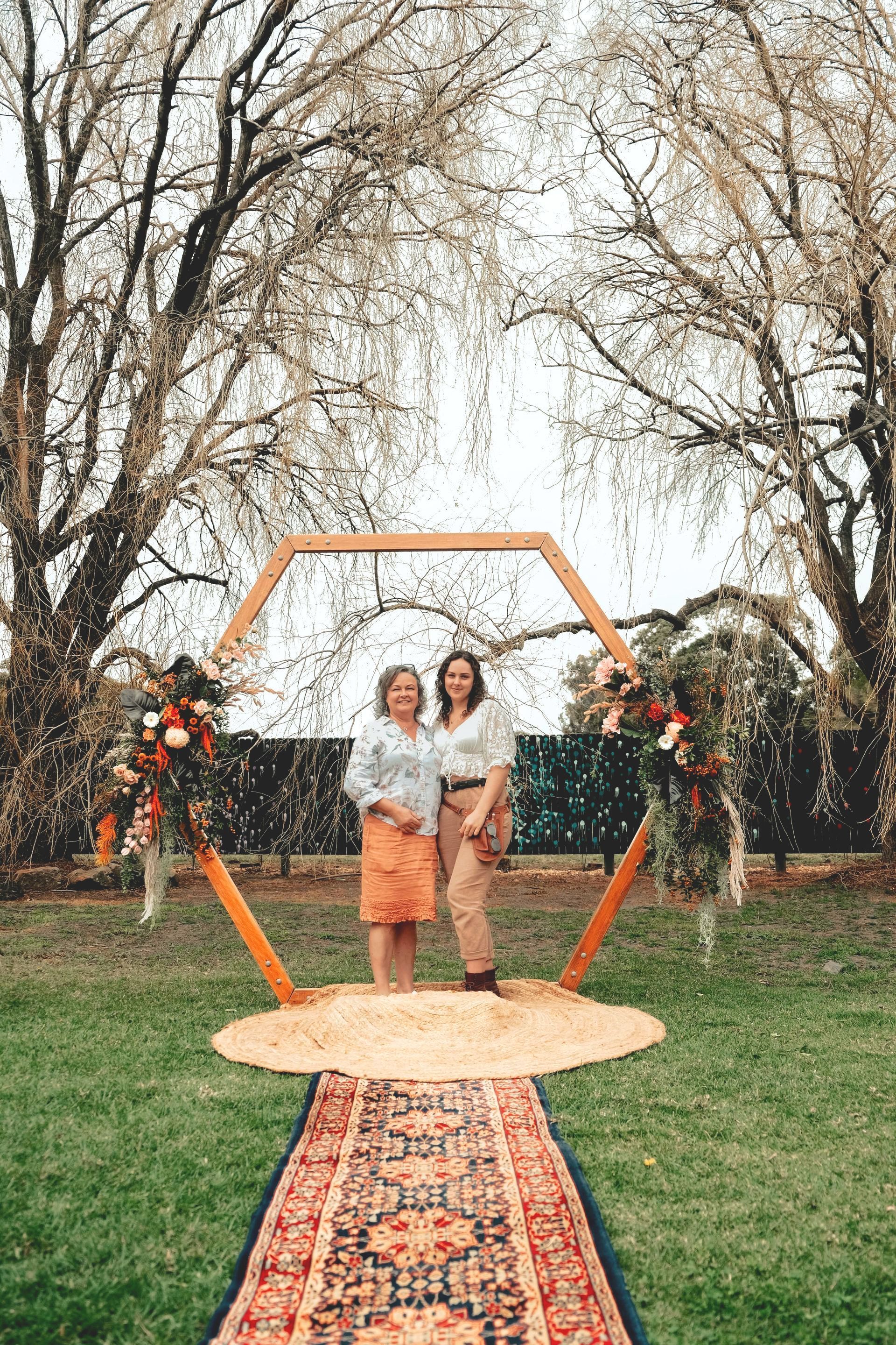Two women pose under a hexagon arch with flowers on a rug in a yard.