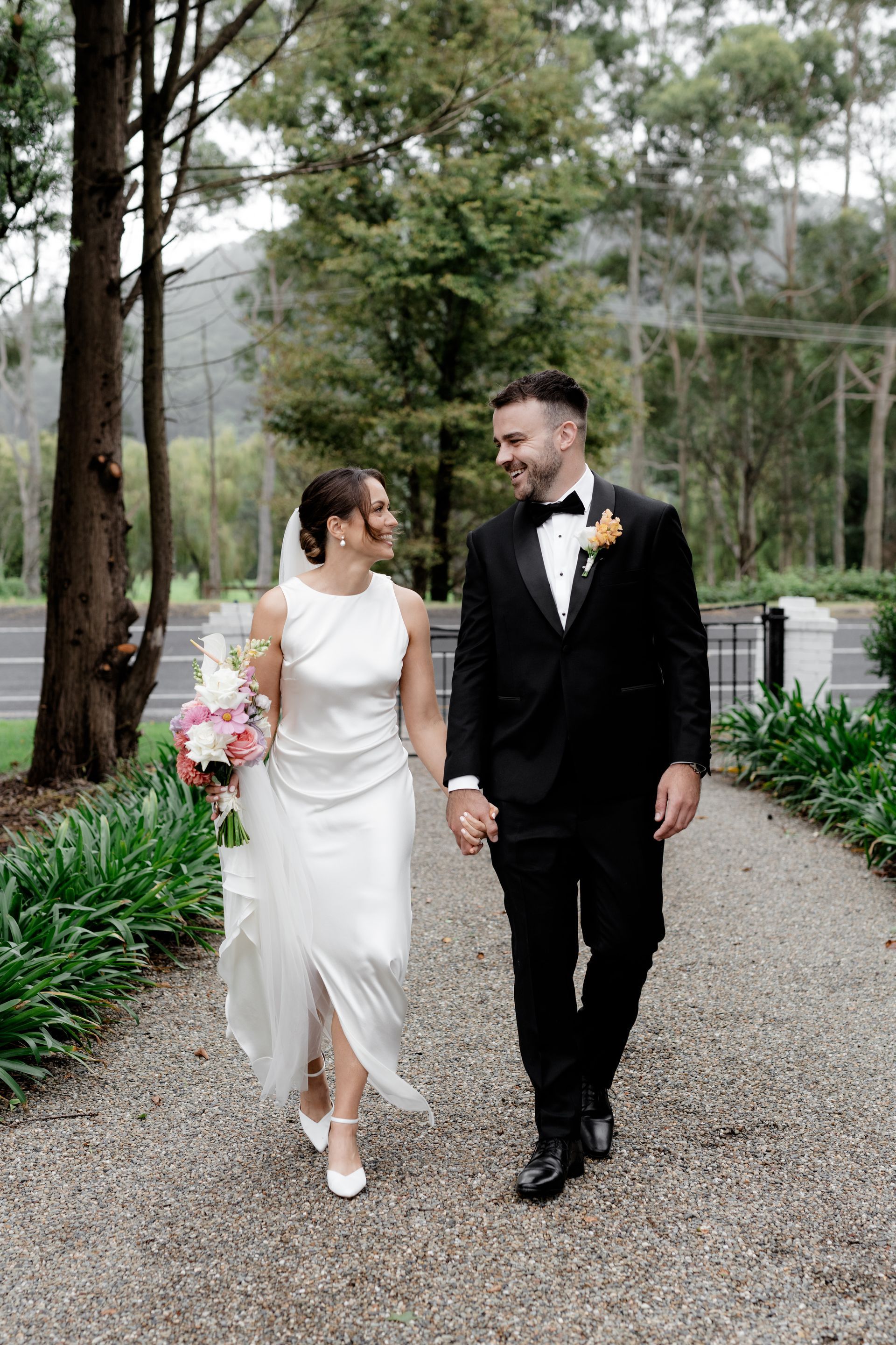 Wedding couple holding hands, walking on a path, smiling at each other. Green trees in the background.