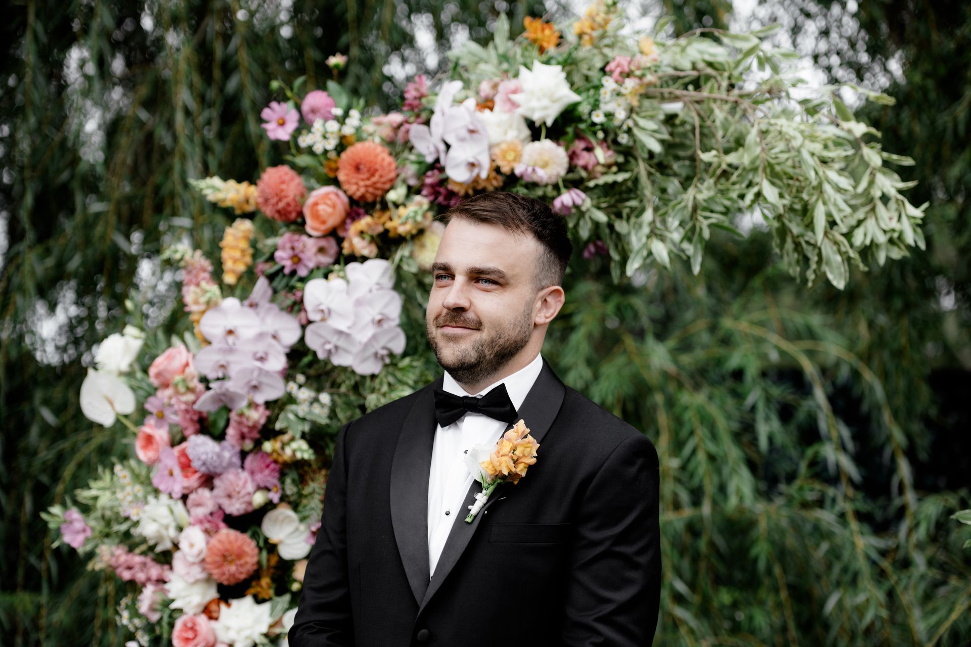 Groom in black tux smiles, awaiting bride. Floral archway backdrop with colorful flowers, outdoor setting.