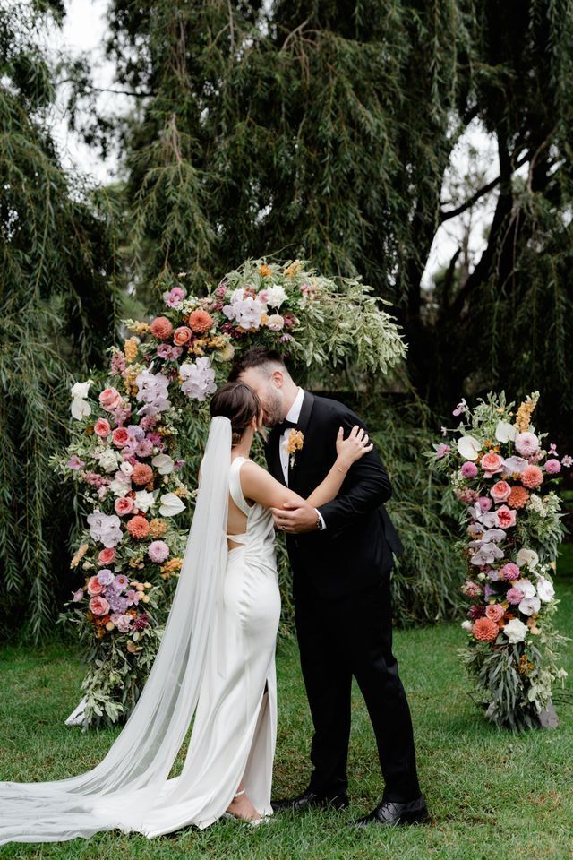 Newlyweds kissing under a floral arch in a garden. Bride in white dress, groom in black suit.