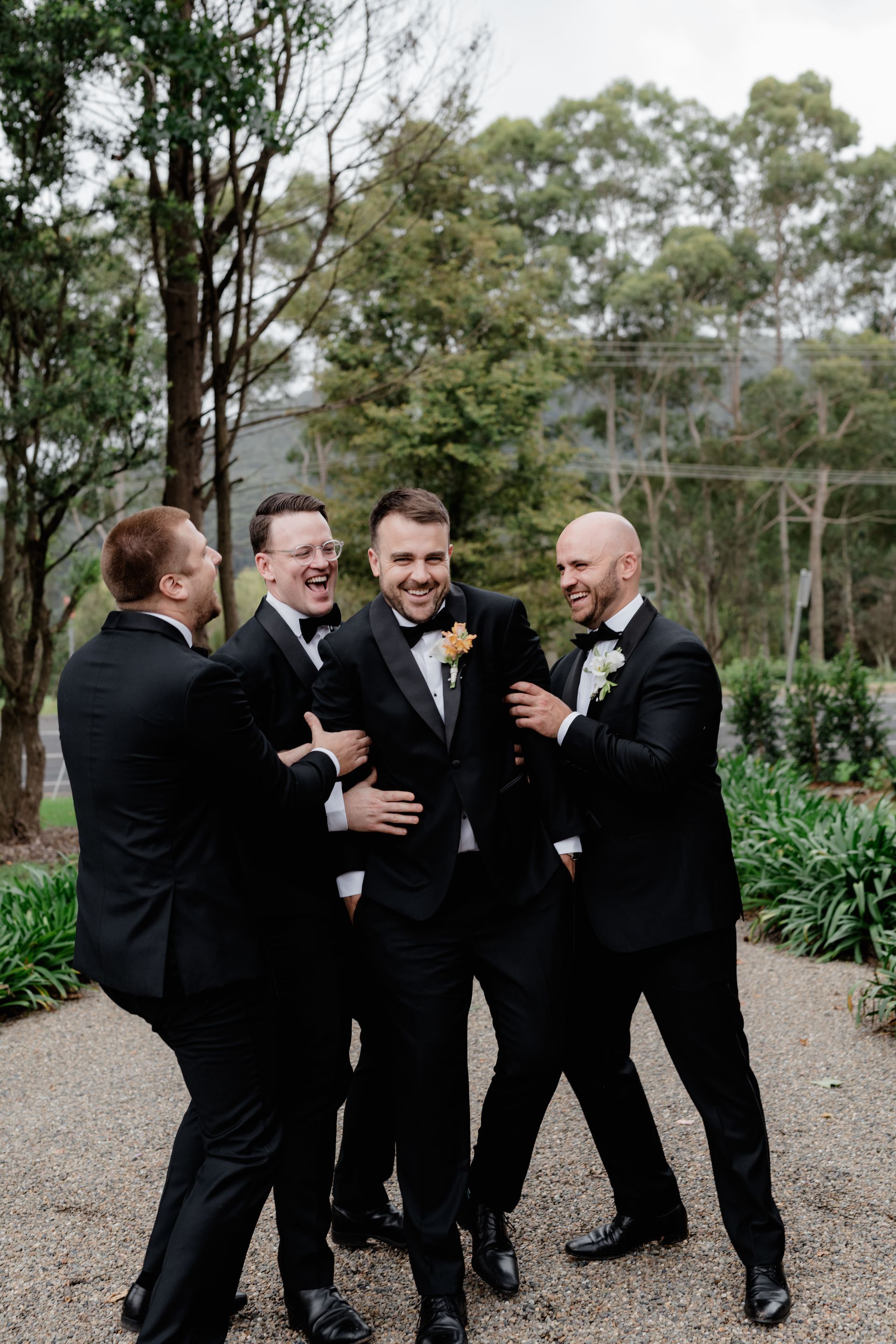 Groom laughing, surrounded by groomsmen in black tuxedos outdoors.