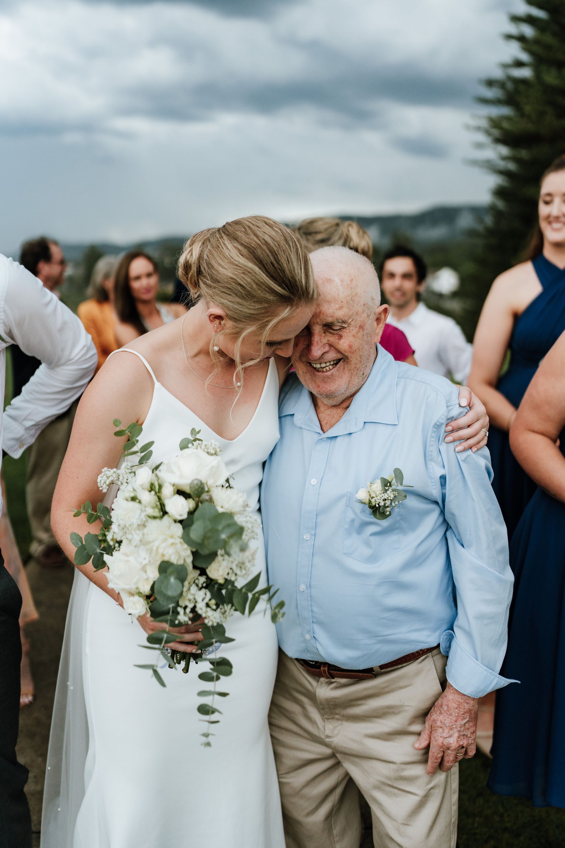 Bride hugs elderly man at an outdoor wedding. She wears a white dress, holding flowers. He wears a blue shirt.