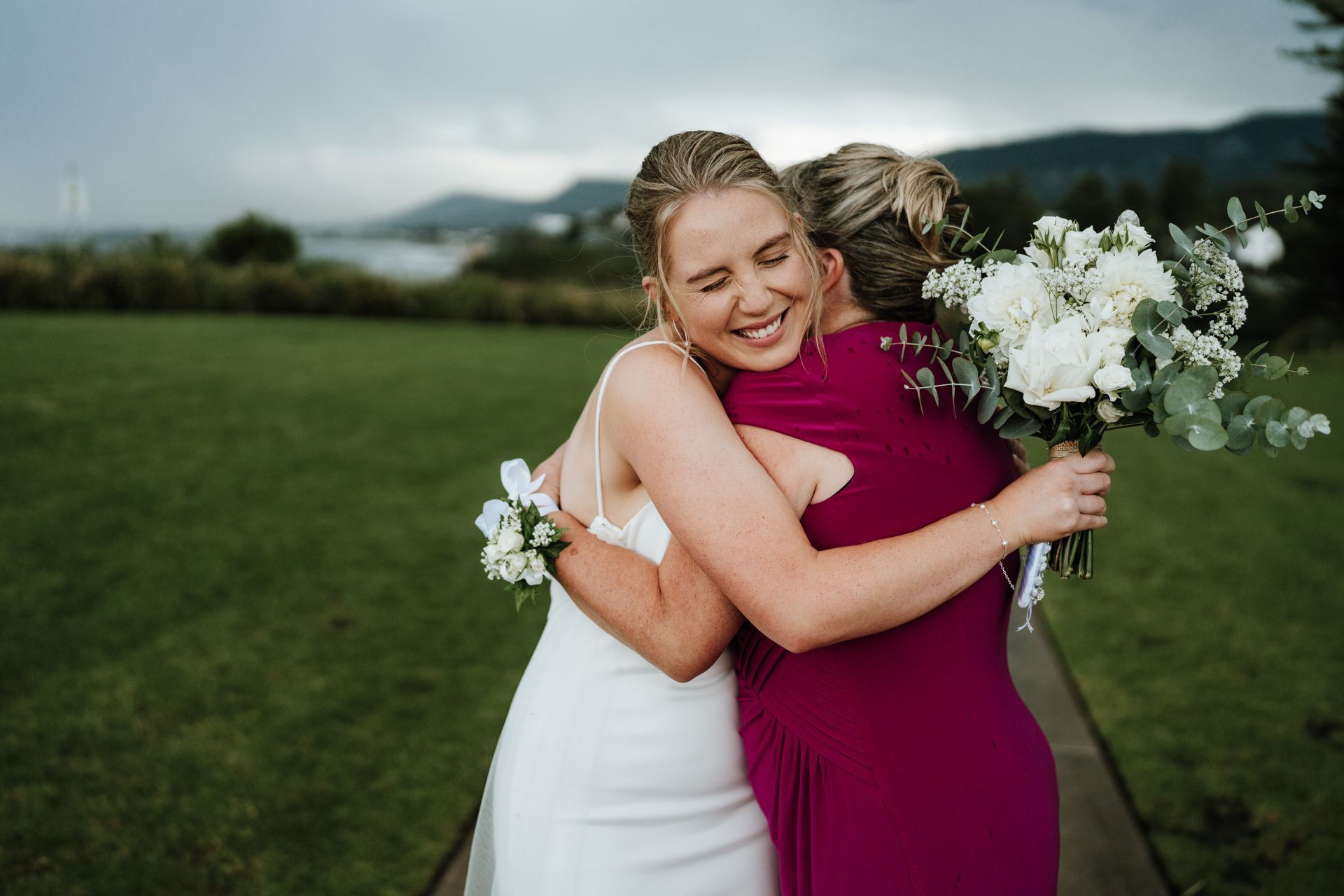 Bride in white dress hugs a woman in magenta dress, holding white flowers, on a green lawn, smiling.