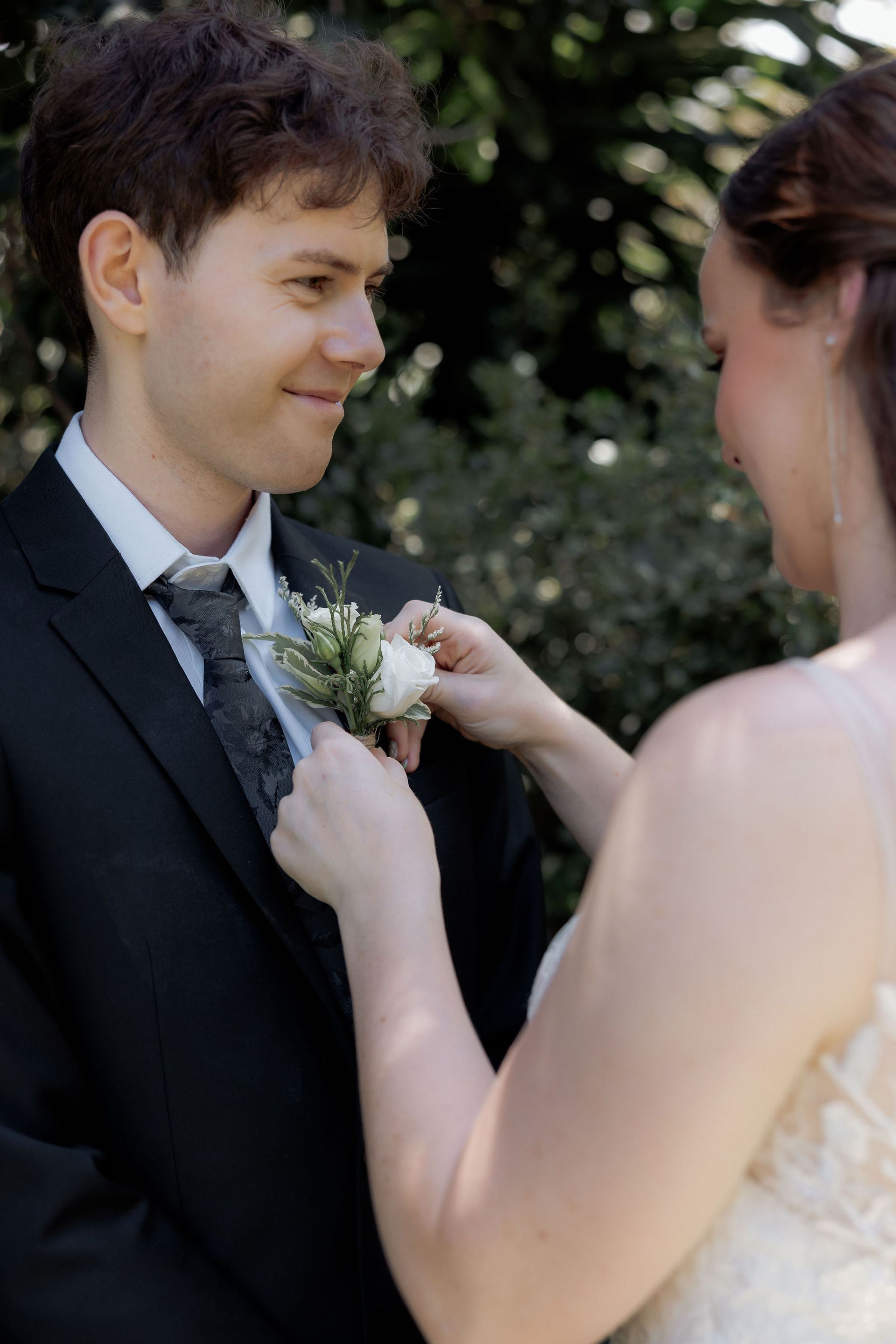 Bride pinning a boutonniere on the groom's suit in a sunny garden, both smiling.