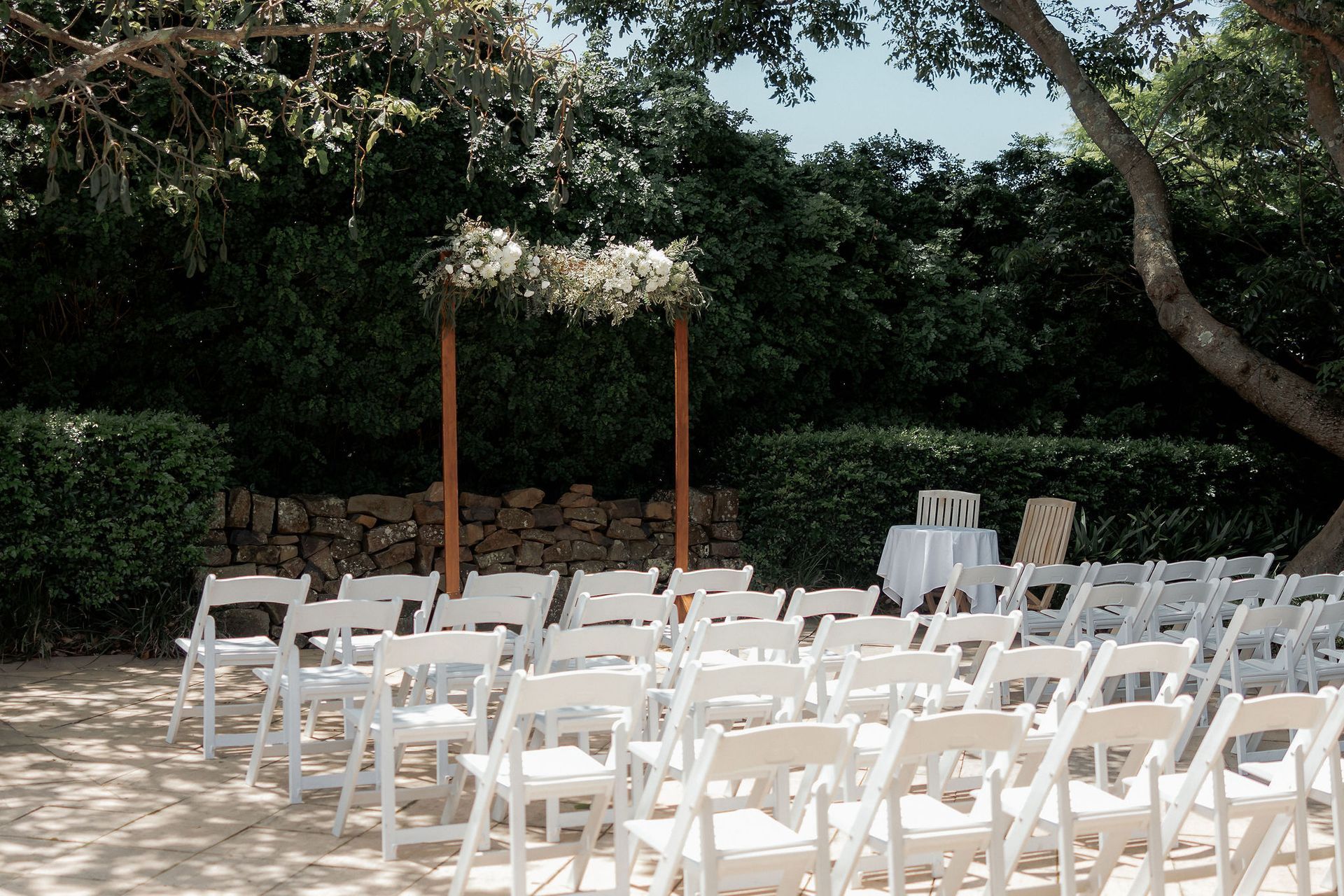 White chairs arranged for an outdoor wedding ceremony; wooden arch with floral decorations; green foliage background.